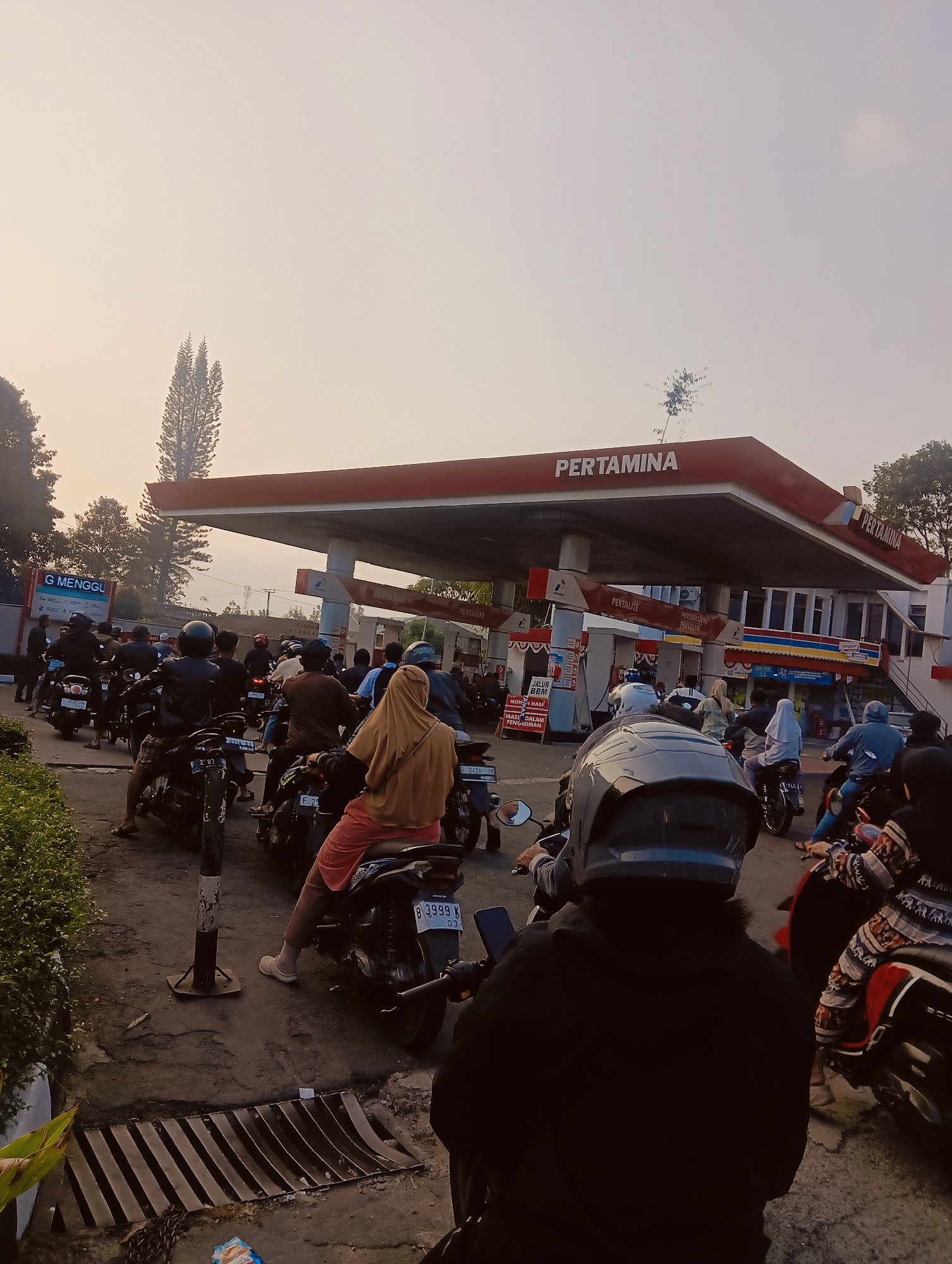 A crowd of people on motorcycles are gathered at a Pertamina gas station in Cisarua, Indonesia. The scene depicts a long queue of motorcycles and riders, many wearing helmets and casual clothing, waiting for service. The gas station has a red roof with the Pertamina logo prominently displayed. Several gas pumps are visible, with some displaying information about fuel types like "Pertamax" and "Pertalite". Signs are also present, including one with the text "JALUR BBM" and "MOHON MAAF MASUK DALAM PENGIRIMAN" (translation: "Fuel Line" and "Sorry, entry in delivery"). The environment suggests an outdoor setting, possibly in the morning or evening given the soft light and haze in the background, which features trees and buildings. Some motorcycles are parked or moving slowly within the queue. The overall impression is one of a busy refueling stop.