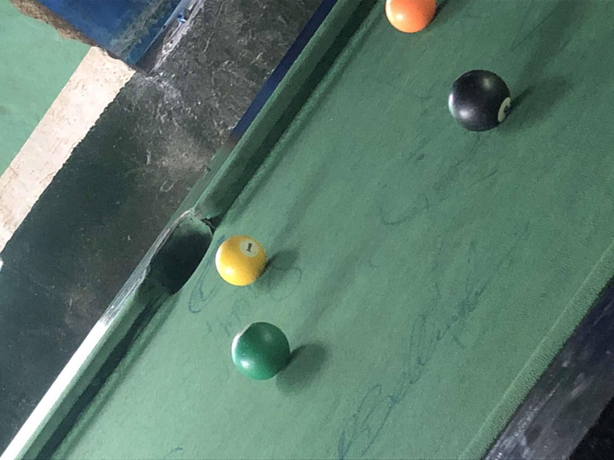 A close-up, high-angle shot of a green billiard table, likely in a casual, possibly outdoor setting in New Stadium, Nigeria, shows four billiard balls. In the foreground, a black eight ball is positioned on the right side of the table. Moving towards the background, an orange ball is visible on the far right, followed by a yellow ball with the number "1" prominently displayed. To the left of the yellow ball is a solid green ball. The table surface appears worn, with some faint blue markings. The background reveals a rough, possibly concrete or stone texture in dark gray and a hint of a blue structure.