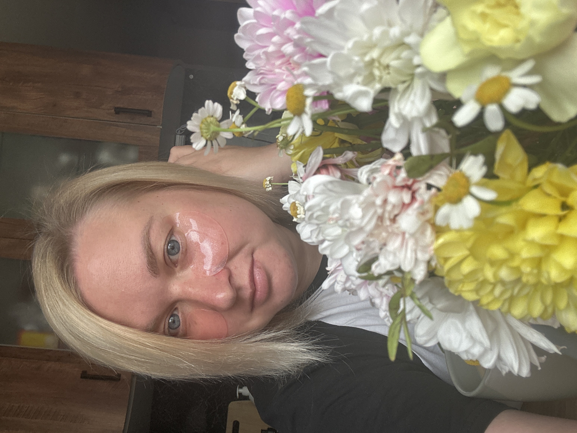 A woman with blonde hair and blue eyes is in a close-up shot, holding a bouquet of flowers. She is wearing a black and white shirt and has pink under-eye masks applied. The background appears to be an indoor setting with wooden cabinets. The image is likely a personal photo taken in a home environment. There is no identifiable information to suggest the location is the city of Chaikovsky, Russia.
