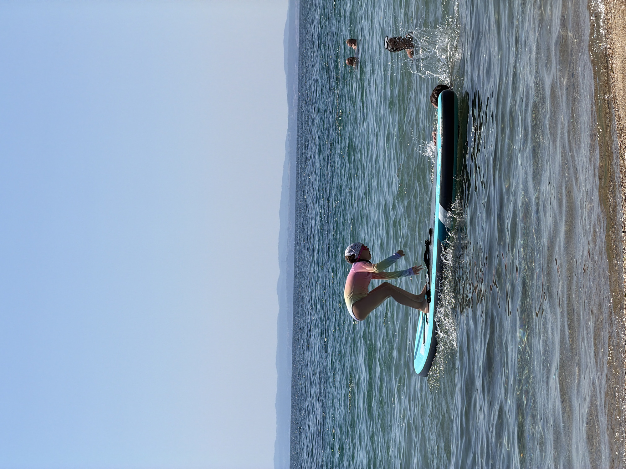 A young girl in a colorful swimsuit and a headscarf is stand-up paddleboarding on a bright blue paddleboard in the Aegean Sea, off the coast of Kalyves Polygyrou, Greece. The water is clear and blue, with gentle ripples reflecting the bright sun. In the background, distant, hazy mountains form a horizon line against a clear, pale blue sky. Several other people are visible in the water further out, some swimming and one person's head is just above the surface, with a splash of water indicating activity. The sandy shore is visible in the foreground, with small pebbles and shells. The girl is in a crouched position, appearing to be adjusting something on the paddleboard, with one hand reaching towards the front of the board. The scene captures a moment of recreational activity on a sunny day at a coastal location.