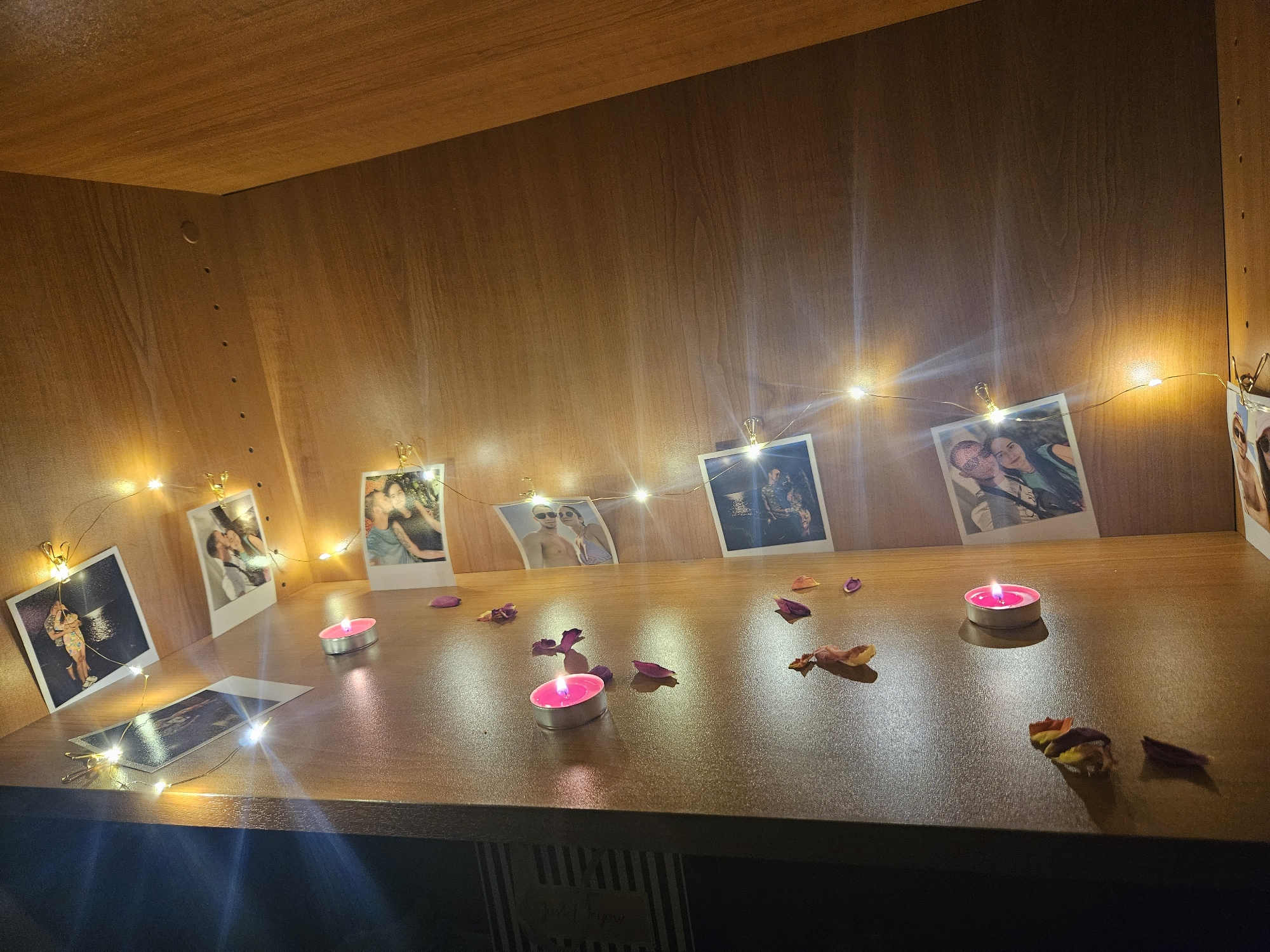 The image depicts a wooden shelf with several framed photographs clipped to a string of fairy lights. The photographs appear to be of couples and are arranged in a somewhat scattered pattern across the shelf. There are also a few lit tea candles and scattered rose petals on the shelf, suggesting a romantic or commemorative display. The background is a plain wooden surface, and the overall lighting is dim, with the fairy lights and candles providing illumination. The location is not specified in the image or its surrounding context.