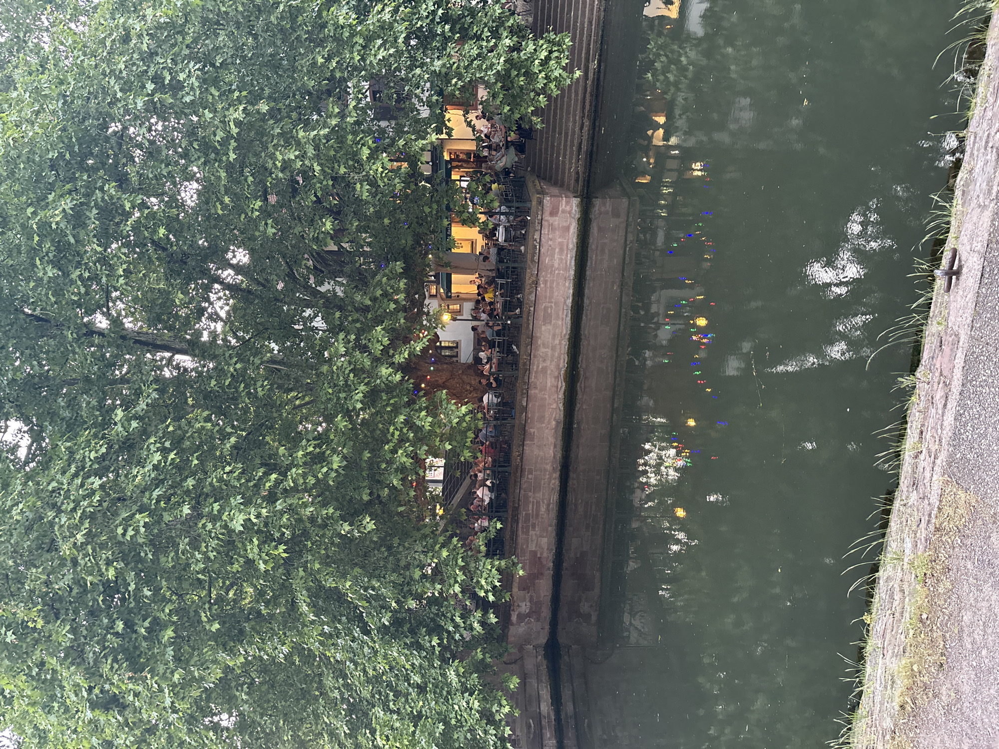The image depicts a canal-side scene in Strasbourg, France. On the left, a dense canopy of trees partially obscures a restaurant or cafe situated along the canal's edge. Patrons are seated at tables, suggesting an active outdoor dining area. The building has a light-colored facade with illuminated windows and awnings. A brick or stone embankment separates the cafe from the canal. The water reflects the lights and colors of the buildings and decorations across the canal. On the right is the concrete edge of a sidewalk. Grass grows in the cracks between the sidewalk and wall.