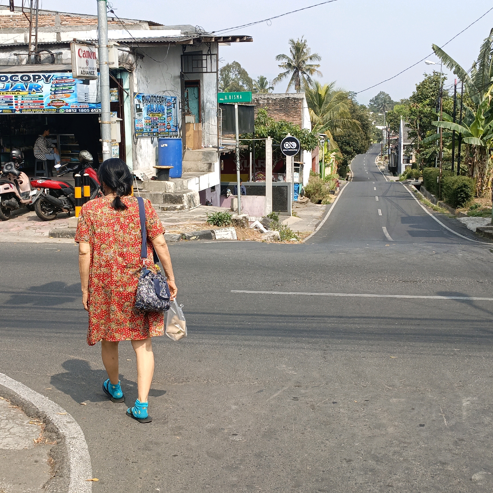 The image captures a street scene in Salatiga, Indonesia, depicting a woman walking away from the camera on a paved road. She is wearing a red and white floral patterned dress, blue socks, and blue sandals. A blue and white patterned bag is slung over her shoulder, and she is carrying a transparent plastic bag in her right hand.
The environment is urban, with buildings lining the left side of the road. One building is a shop with signage including "Canon" and "PHOTOCOPY FAJAR," suggesting it is a copy and printing service. A person is seated inside the shop. Motorcycles are parked in front of the shop. A green street sign with "JL. BISMA" is visible on the left side of the road.
The road itself is asphalt, with white lane markings. Trees and vegetation are present on both sides of the road, with palm trees visible in the background. The sky is clear and bright, indicating a sunny day. The scene suggests a typical day in a residential or commercial area of Salatiga.