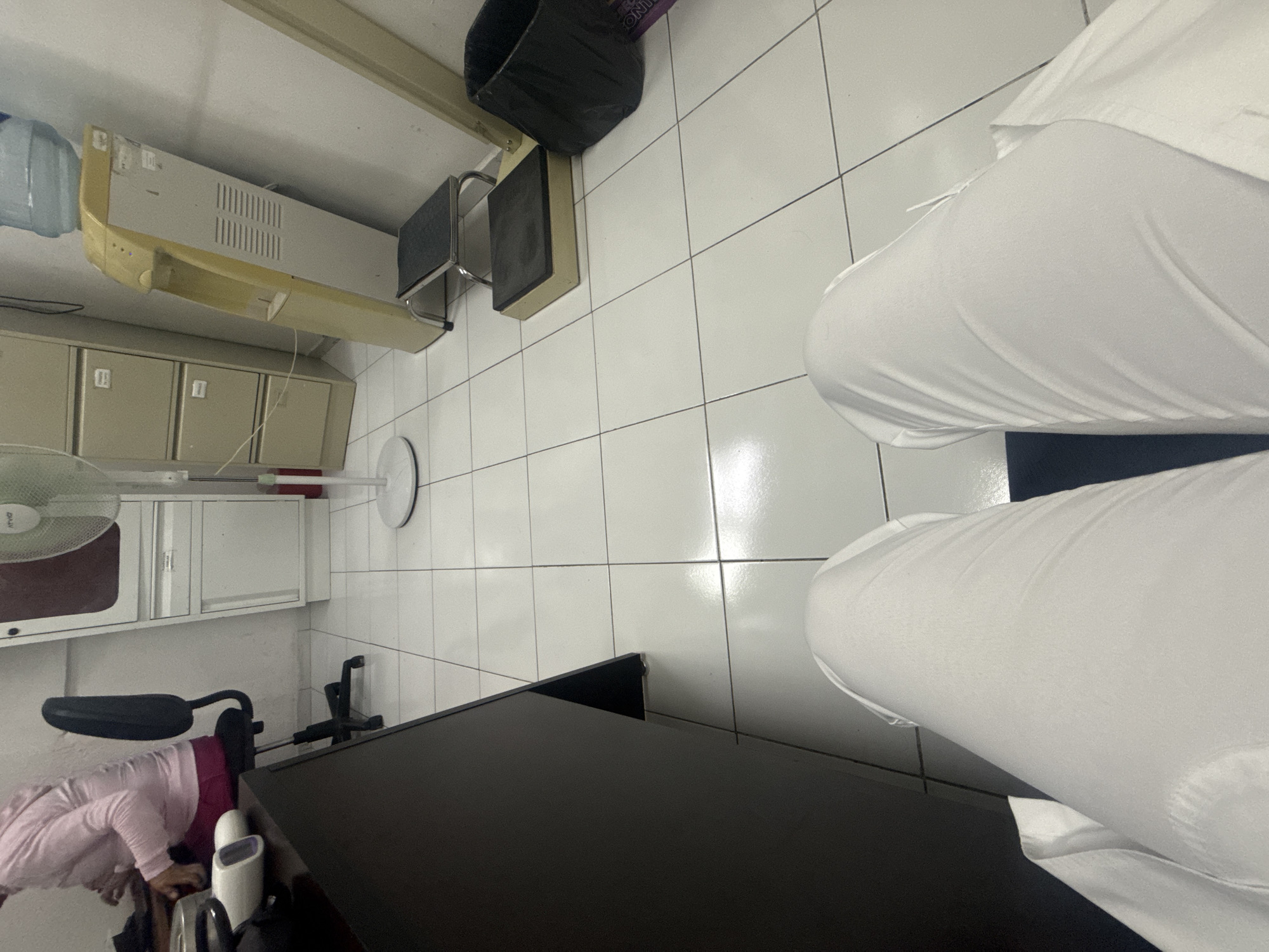 A high-angle shot depicts a portion of a room with white tiled floors. The room contains a black trash can, a footstool, and a small cabinet with a sink. A floor fan, along with what appears to be medical equipment, are situated on the left side of the frame. A black table occupies the lower center. A person wearing white clothing is positioned in the upper right of the scene. The image could be from a clinic or hospital environment. The location in Puebla, Mexico, is not determinable from the image.