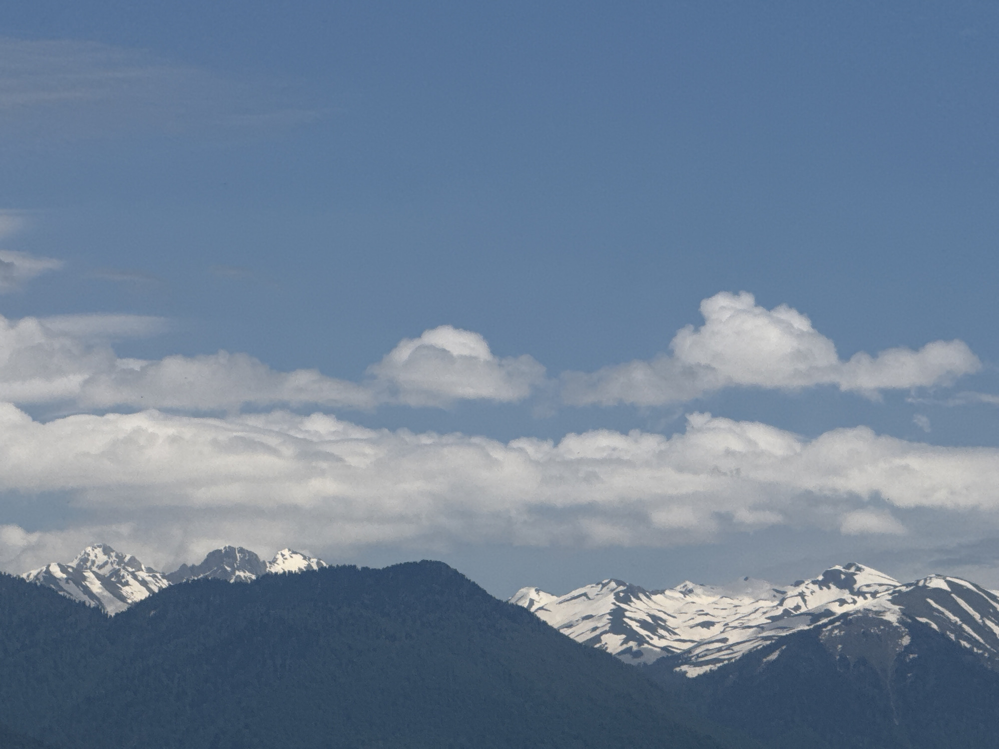 The image depicts a mountain range with snow-capped peaks under a blue sky with scattered white clouds. In the foreground, there are dark green, tree-covered mountains. Above these, there are smaller, snow-covered peaks that appear to be the higher sections of the mountain range. The sky is a clear blue color in the upper portion of the frame, with patches of white clouds at the center. The location is not explicitly clear from the image and cannot be confirmed from the given information.