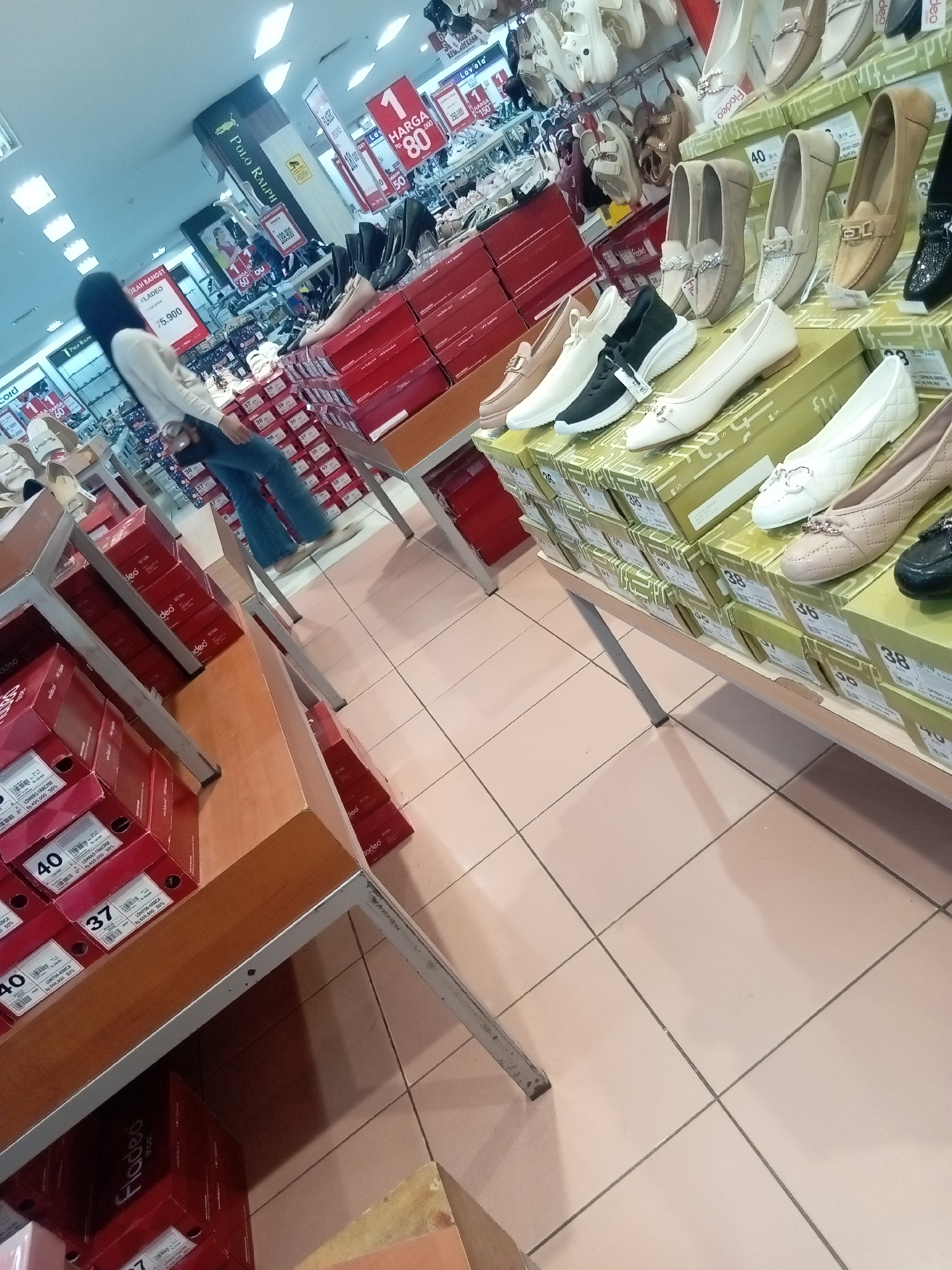 The image depicts the interior of a shoe store in Batam, Indonesia, likely a department store shoe section. A woman wearing a black hijab and a light-colored sweater with blue jeans is walking away from the camera, towards shelves of footwear. The shelves are organized with shoe boxes and displayed shoes. In the background, promotional signs are visible, one advertising "HARGA 1" (Price 1) and "80%" off. The floor is tiled with a light-colored pattern. The overall environment suggests a retail setting with a focus on women's shoes, with a variety of styles and colors displayed on shelves and in boxes.
