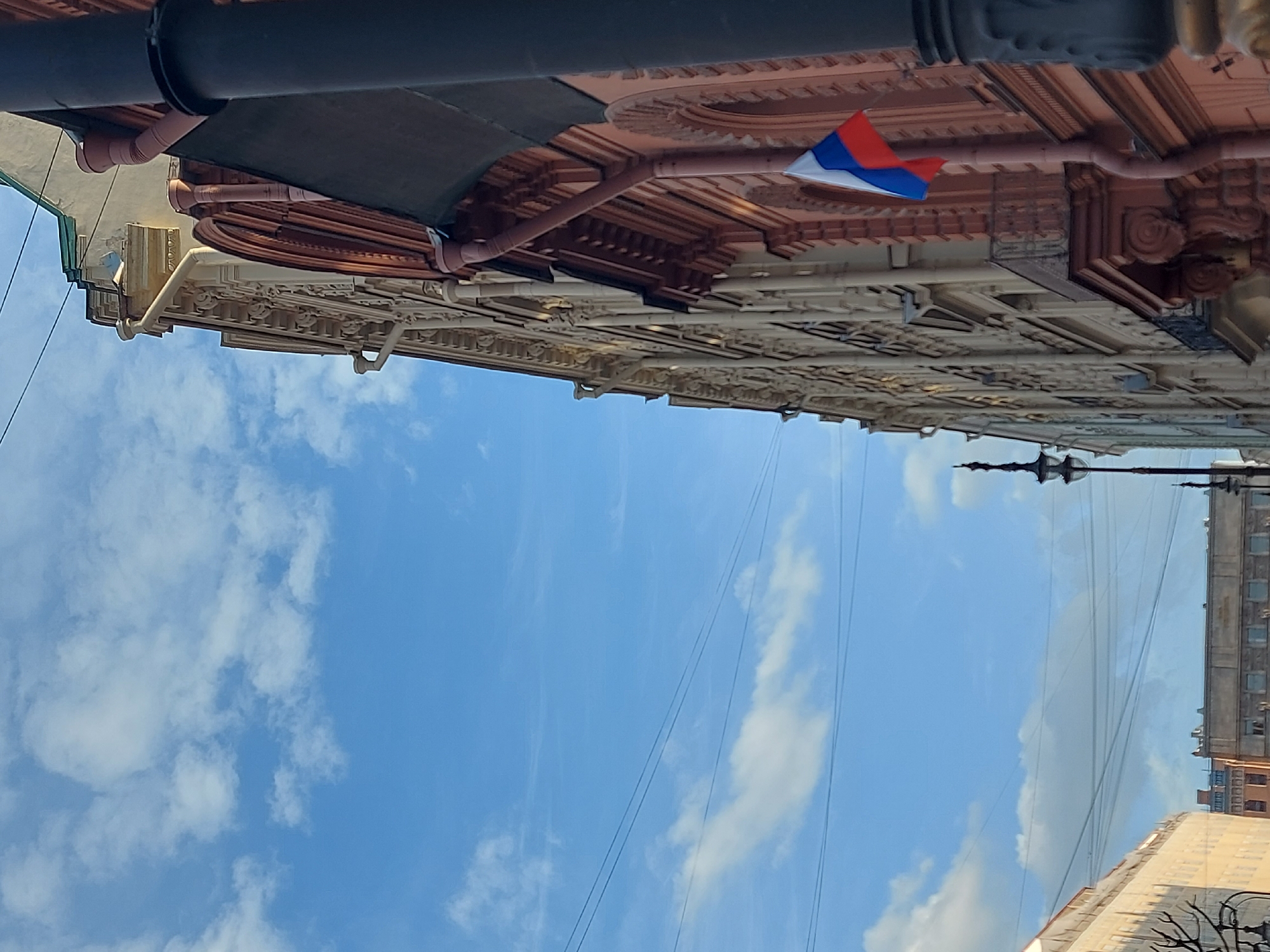 The image depicts a view of a building facade in what appears to be Saint Petersburg, Russia. The architecture is ornate, with detailed cornices, moldings, and decorative elements. A Russian flag is prominently displayed, attached to a pipe or structural element on the building. The sky is visible as the backdrop, featuring light clouds. Power lines are seen against the sky. Other buildings are partially visible in the background.