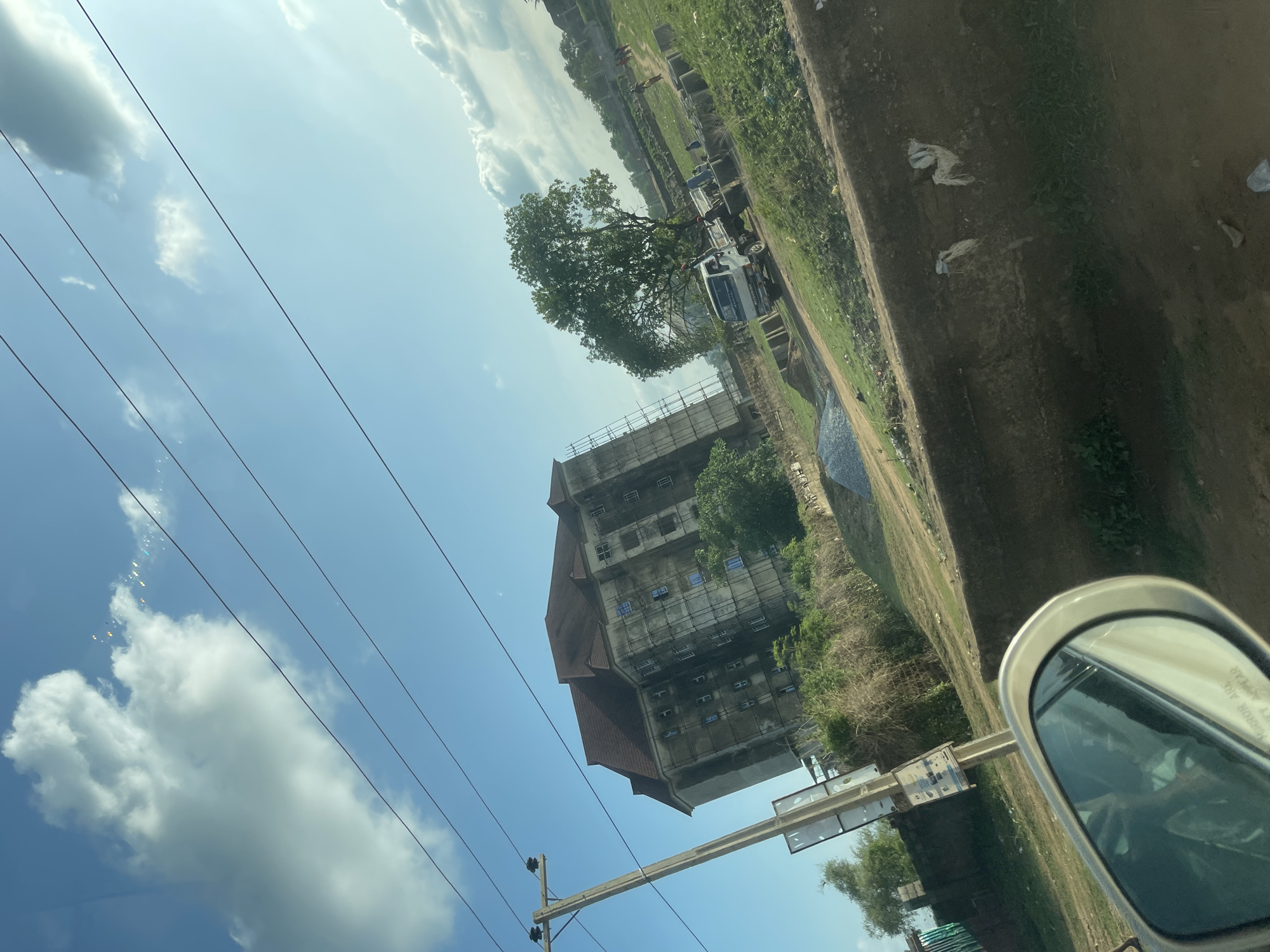 The image shows a partially-constructed building with scaffolding in Abuja, Nigeria. The building has a red roof and appears to be several stories tall. Power lines are visible against a blue sky with clouds. A dirt road runs along the side of the building, and there is some vegetation. A vehicle's side mirror is visible in the foreground. A truck is parked near the construction site. Some people are visible in the far background. Debris is on the roadside.