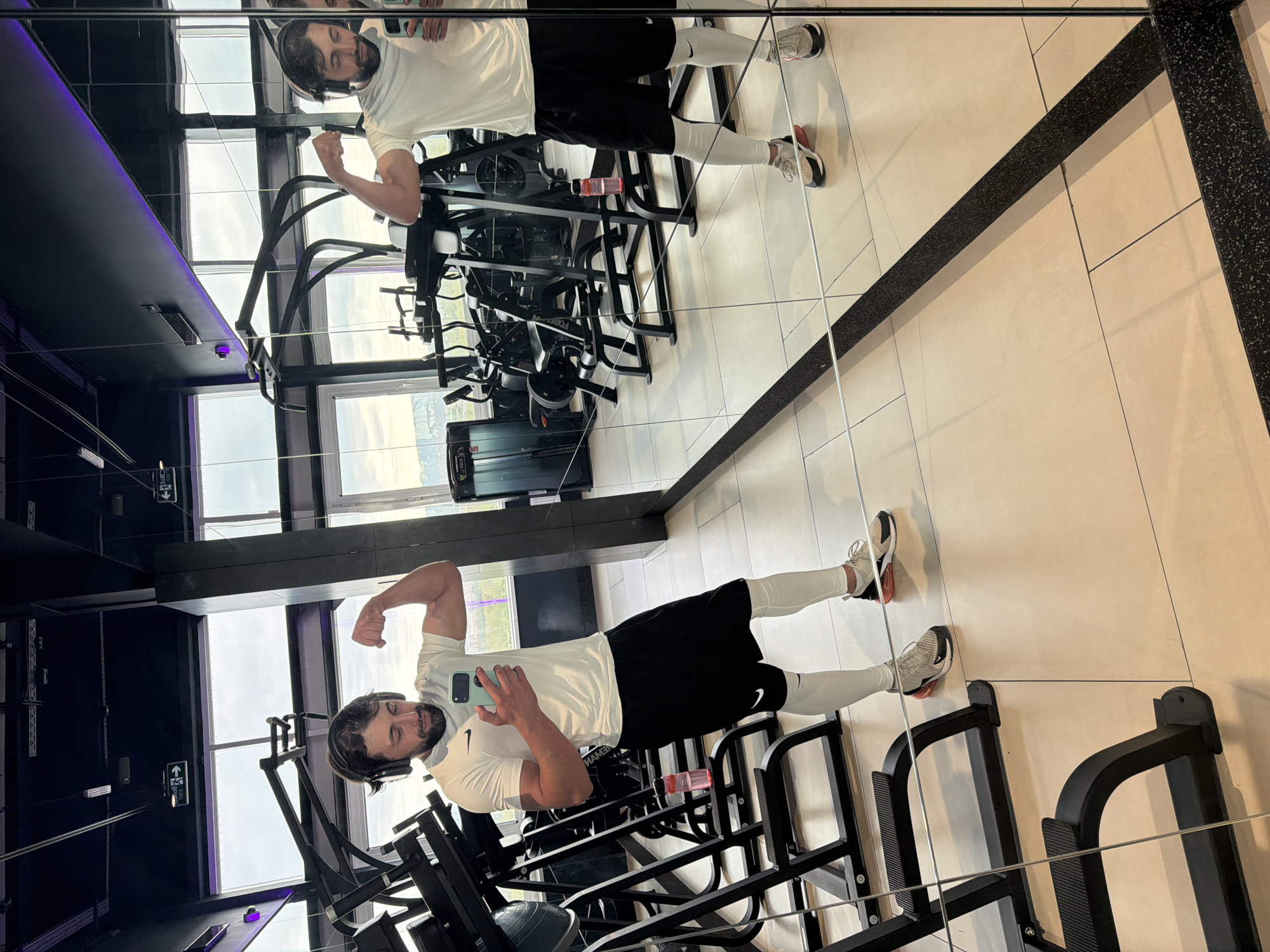 A man with dark hair and a beard, wearing a white t-shirt, black shorts, white leggings, and athletic shoes, is posing for a photo in a gym. He is holding a smartphone in his left hand and flexing his right arm. He is standing in front of a mirror that reflects his image and the gym equipment behind him. The gym appears to have weight machines and a dark ceiling with purple accents. The background shows windows with a view of the sky, suggesting a location at some height. The image was taken indoors. Based on the information provided, it would be surmised that the gym is in Krasnoyarsk, Russia.