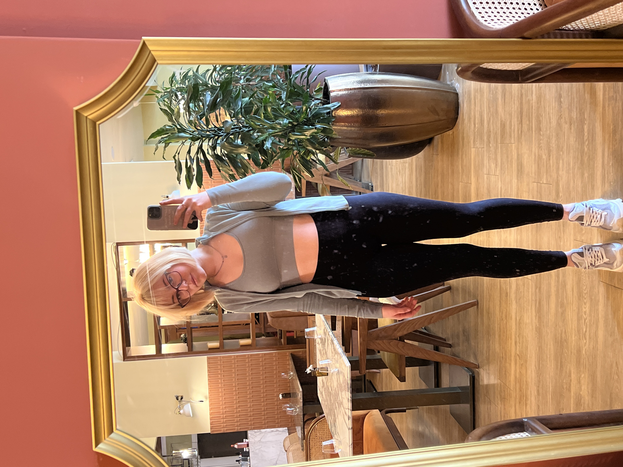 A full-length reflection in a gold-framed mirror of a fair-skinned woman with blonde, shoulder-length hair wearing glasses, a gray cropped tank top, a light gray cardigan, black leggings with a white speckled pattern, and white sneakers. She is holding a cell phone and facing the mirror. The reflection also includes a potted plant, a wooden floor, wooden furniture, and a table with glassware and floral arrangements. The mirror is hung on a salmon-colored wall. The scene appears to be inside a restaurant. Location of this image in Boreti, Montenegro is not possible to ascertain, since it is impossible to tell from the image.