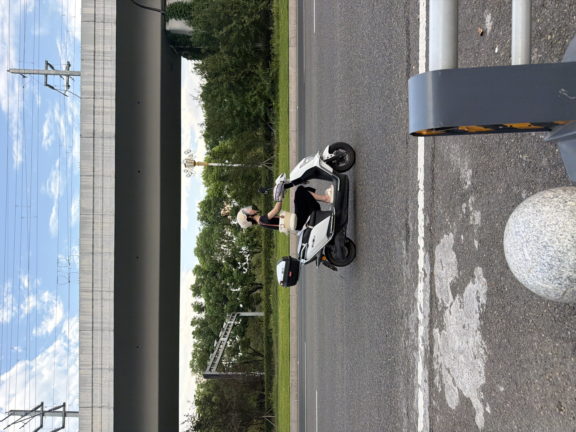 In the city of Jiangjin, China, a woman wearing a white helmet and a black outfit rides a white electric scooter on an asphalt road. She is carrying a white bag over her shoulder and has a black storage box attached to the back of the scooter. The scooter is in motion, slightly angled towards the viewer. In the background, a concrete overpass runs horizontally across the frame, with power lines and a blue sky with clouds above it. Trees line the median of the road and the embankment beyond. The foreground shows a portion of the asphalt road with white lane markings and a grey, rounded bollard and metal barrier.