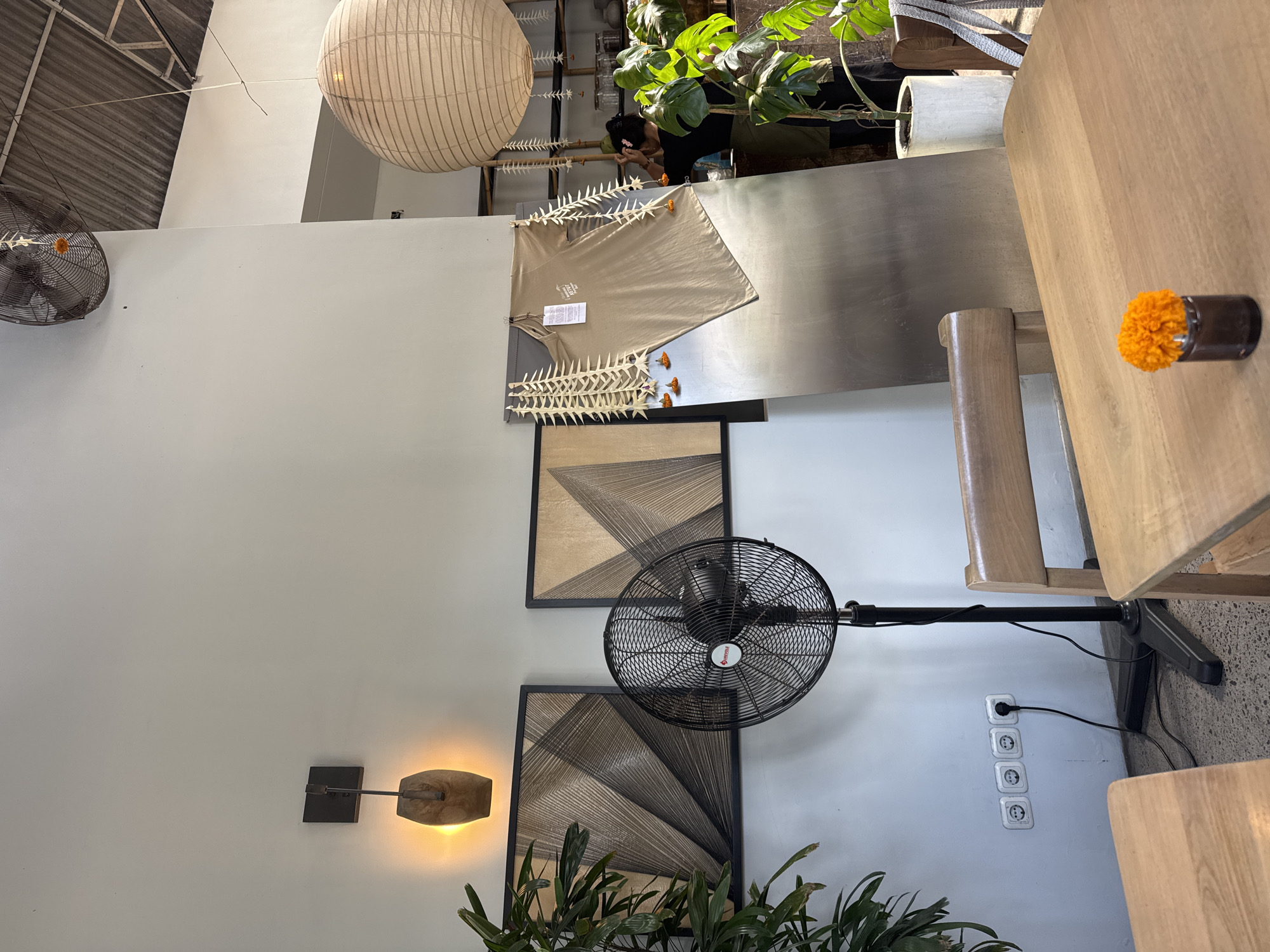 The image depicts the interior of a cafe or shop in Mengwi, Indonesia. The setting appears to be a bright, airy space with white walls and a corrugated metal ceiling. In the foreground, a light wooden table holds a small glass vase with an orange marigold flower. To the left of the table, a portion of another wooden table is visible.
The middle ground of the image is dominated by a black standing fan positioned in front of two framed pieces of art that appear to be geometric abstract designs made of string on a textured background. Above the artwork, a wall-mounted light fixture casts a warm glow. To the right of the fan and artwork, a large, leaning metallic sheet serves as a backdrop for a tan t-shirt hanging on display, adorned with decorative white palm frond ornaments and small orange flowers.
In the background, a large, spherical paper lantern hangs from the ceiling. A woman, seen from the back, is partially visible on the right, interacting with a large potted plant with broad leaves. Another smaller plant is visible in the left foreground. Electrical outlets are visible on the lower portion of the wall near the fan. The floor is concrete.