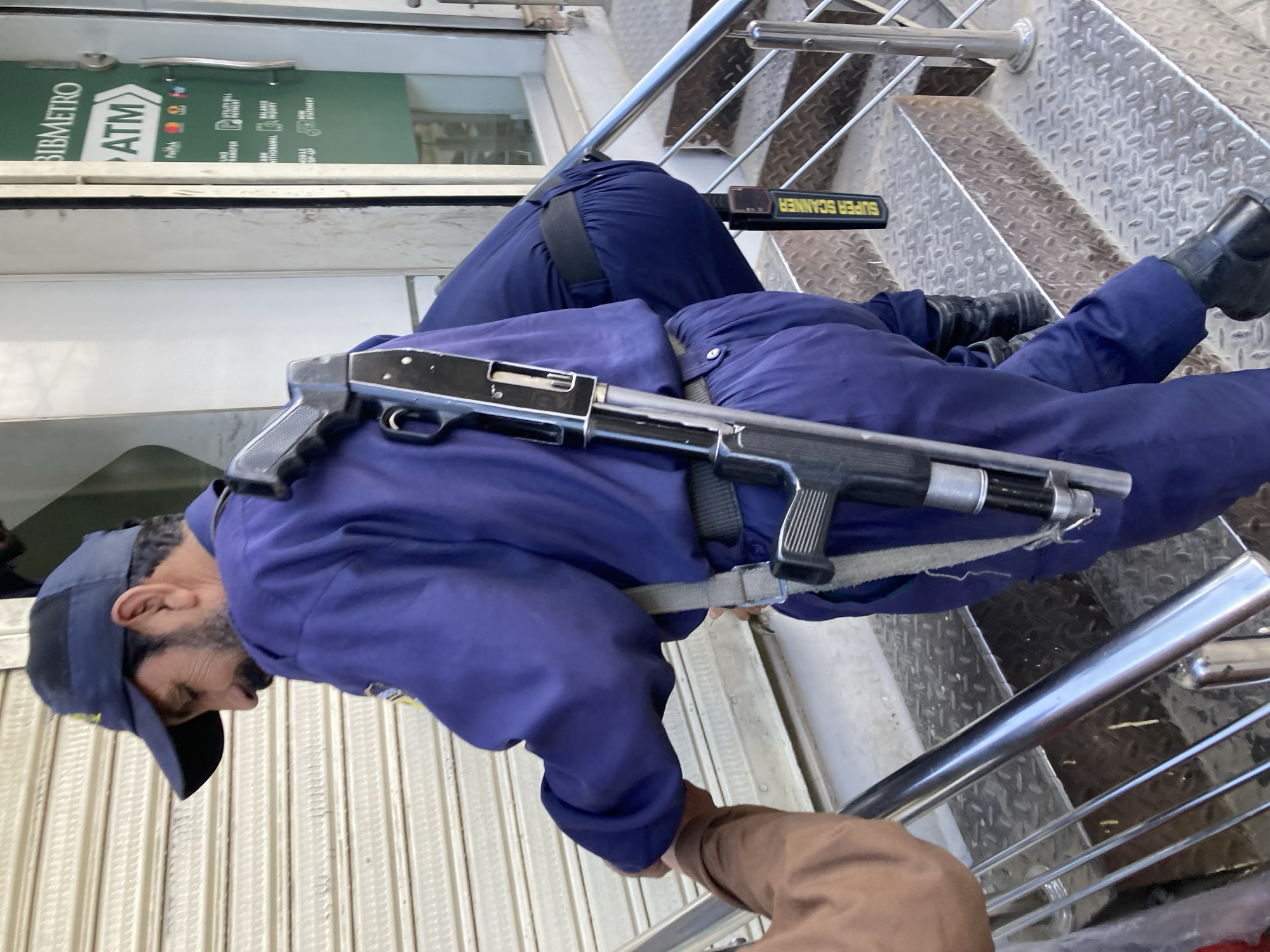 A man in a navy blue uniform and cap is standing on a metal staircase, holding a shotgun. He also has a "SUPER SCANNER" device attached to his belt. The "BIBMETRO ATM" is visible in the background. The location is Quetta City Tehsil, Pakistan.