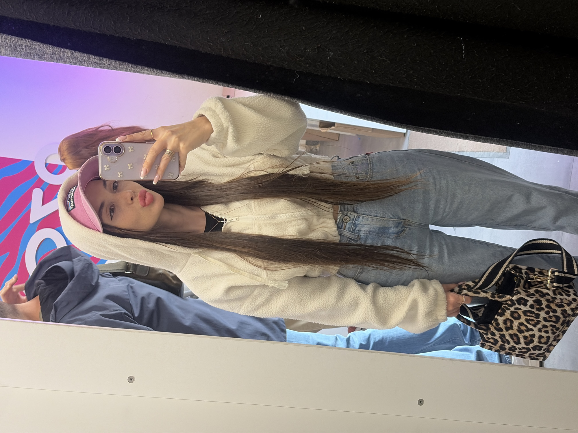 A young woman is taking a selfie in what appears to be a dressing room or a similar retail space. The woman has long, straight brown hair, and she is wearing a pink cap, a white, fluffy hooded jacket, and blue jeans. She holds up a smartphone with a decorated case. Her lips are pursed in a kissing expression. To her left is the reflection of a man in a blue jacket. A patterned handbag sits to her right. Behind her, there's a colorful design of blue, pink, and white. The location is not identifiable as Saint Petersburg, Russia, from the visible elements in the image.
