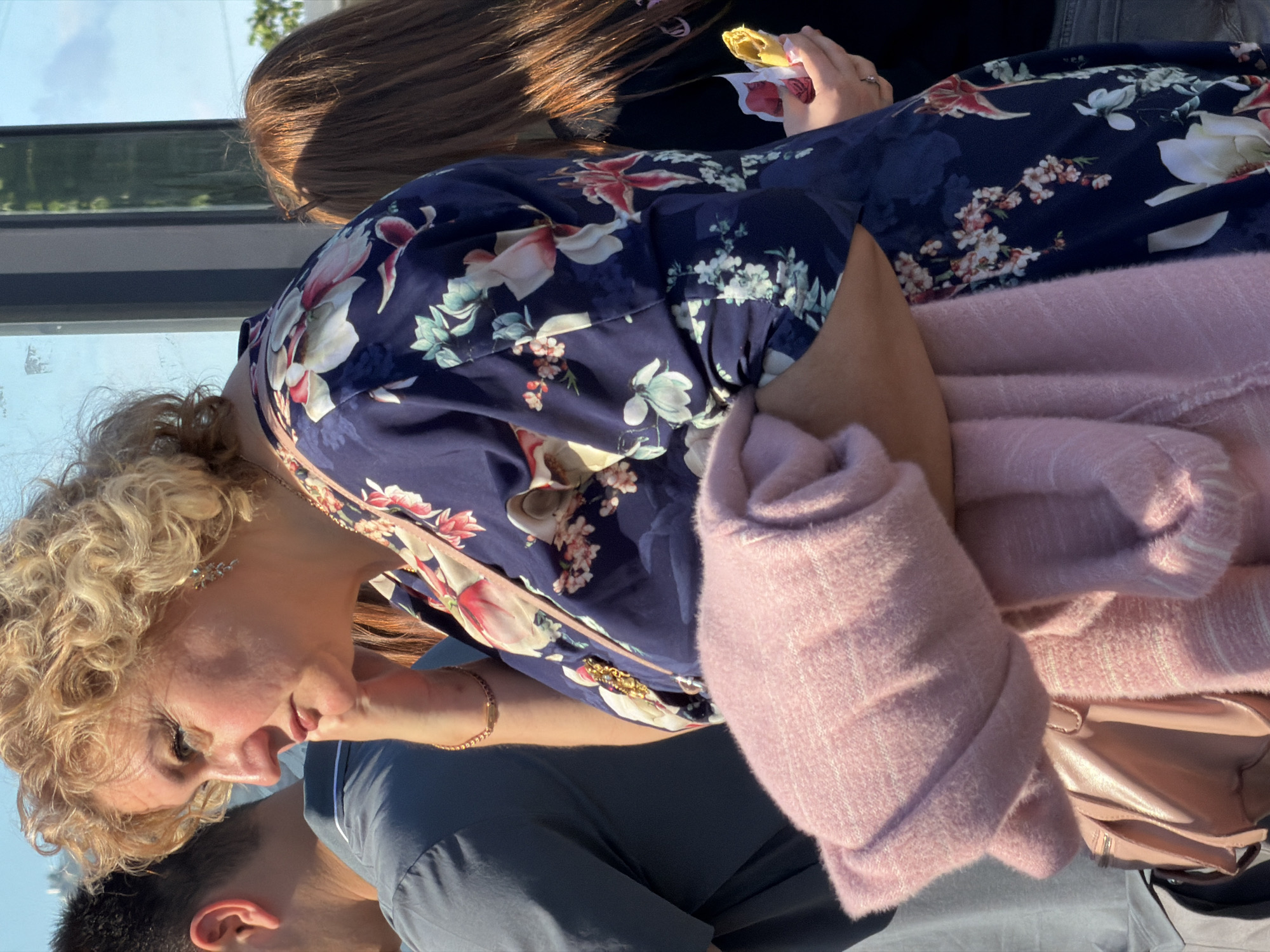The image captures a casual social gathering, primarily featuring a woman with curly blonde hair in profile, looking downwards towards another individual whose dark-haired head is visible in the bottom left. She wears a navy blue dress adorned with a vibrant floral pattern of white, pink, and green blooms. A soft, light pink, fuzzy sweater or shawl is draped over her right arm and shoulder, partially obscuring a light pink handbag.
Behind her, the upper part of another person with long brown hair is visible, holding a yellow pastry-like food item wrapped in white and red paper. The blonde woman has a gold-colored bracelet on her left wrist and a delicate, silver-toned earring.
The scene is brightly lit by natural sunlight, suggesting daytime with clear weather, as indicated by the reflections of a bright sky and trees visible through a window or glass barrier in the background. The overall mood is relaxed. No visible text is present in the image. The provided context indicates the location is Saint Petersburg, Russia.