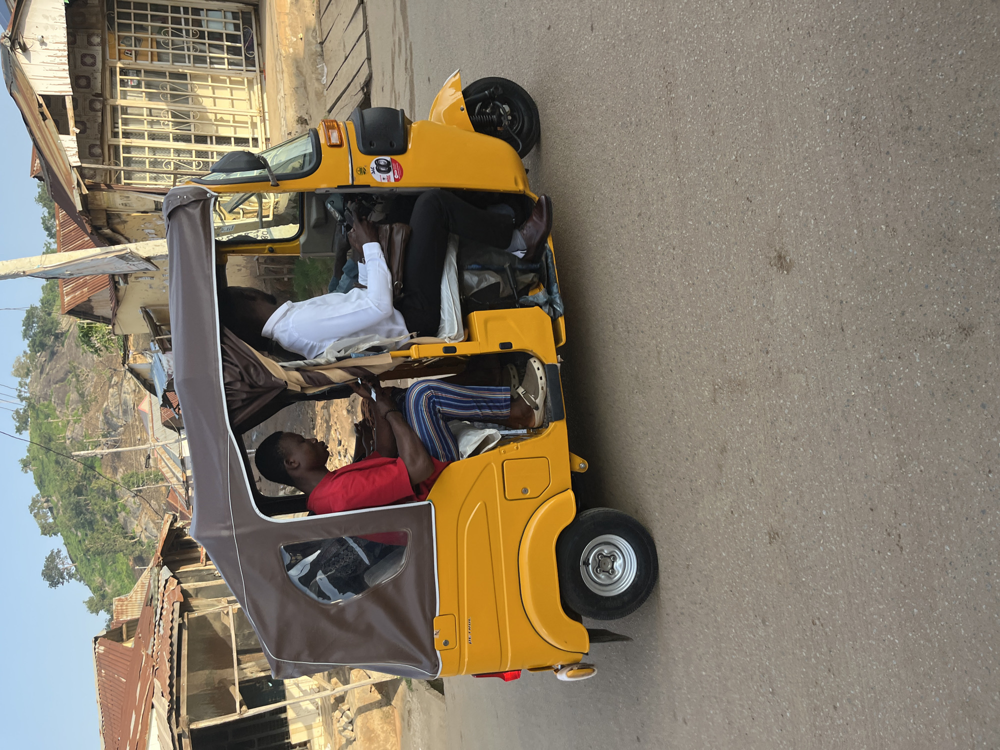 In Abuja, Nigeria, a yellow auto-rickshaw, a common mode of transportation, travels on a paved road. The auto-rickshaw, also known as an "Keke Napep," carries two occupants. The driver, wearing a white collared shirt and black trousers, operates the vehicle. In the passenger seat behind him, a man in a red shirt and striped trousers is seated, looking down at his phone. The background reveals a neighborhood with buildings featuring corrugated iron roofs, characteristic of some areas in Abuja, and a rocky, tree-covered hill. The sky is clear and bright, suggesting daylight.