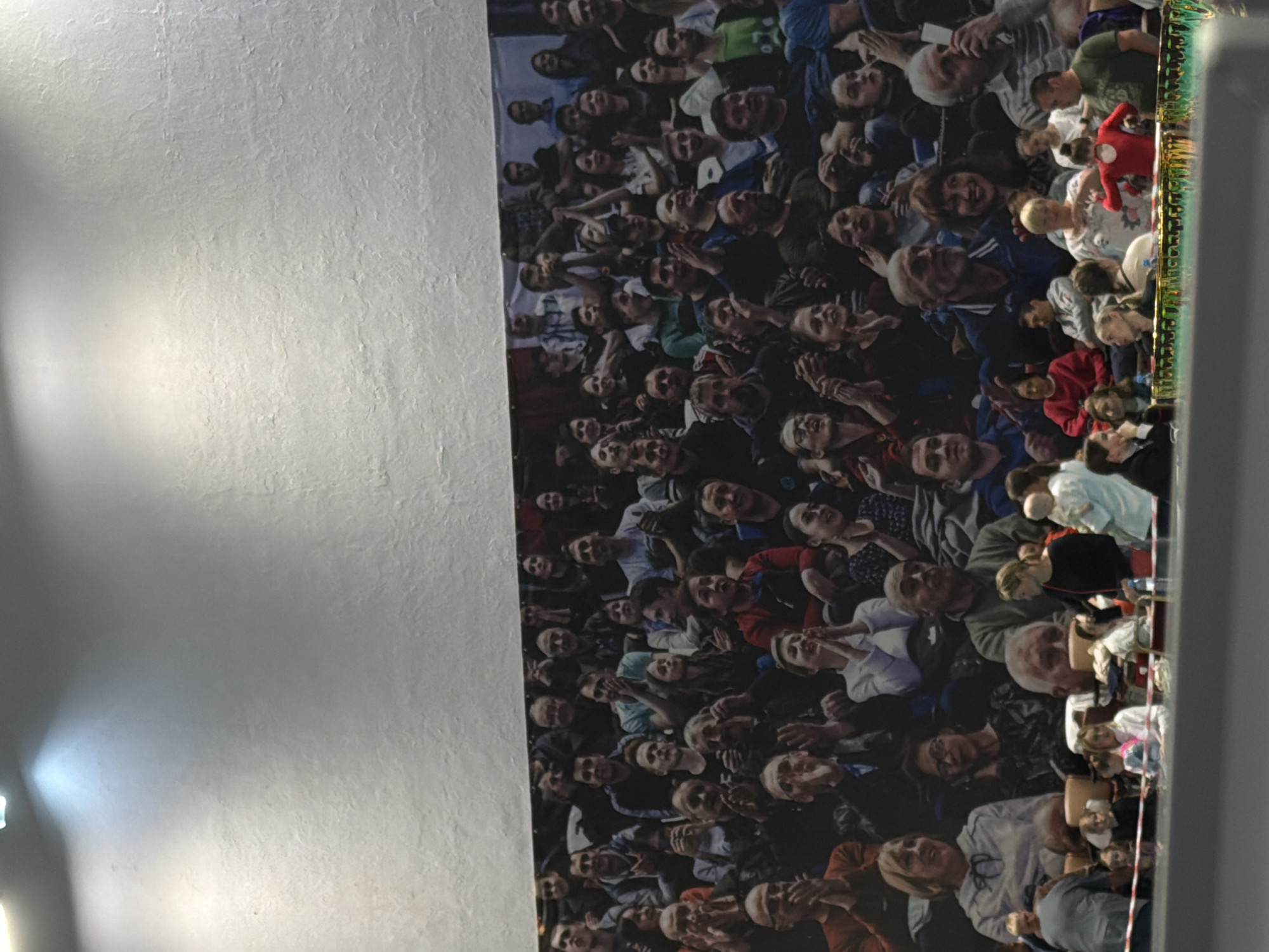 This image depicts an indoor scene in the city of Perm, Russia, featuring a large wall mural of a crowd above a section of real-life spectators.
The upper portion of the image shows a light grey, textured wall, possibly stucco or plaster, illuminated by two distinct, cool-toned overhead lights that cast bright beams downwards. Below this, a large, rectangular banner or mural covers the majority of the wall, depicting a dense, multi-ethnic crowd of individuals, predominantly adults, seated in what appears to be an arena or auditorium. Many of the depicted figures are shown clapping, smiling, or observing an unseen event. Visible within the depicted crowd, near the upper right corner of the mural, is a flag with two horizontal stripes, red over white, resembling the flag of Poland.
In the foreground, below the mural, real individuals are present, observing an event that is out of frame. This group includes adults and several children. A woman in a white and red patterned top stands, holding a baby. Another individual, possibly a child, is visible in a bright red leotard, indicating a potential performance or sporting event. People are seated in rows, some facing left and others right from the viewer's perspective. Several individuals in the foreground are engaged in conversation or observation. A distinctive green and gold ornate railing or barrier separates the immediate foreground from the main area of activity, with a white and red striped tape barrier also visible closer to the viewer. The overall lighting is bright and artificial, consistent with an indoor public venue.
