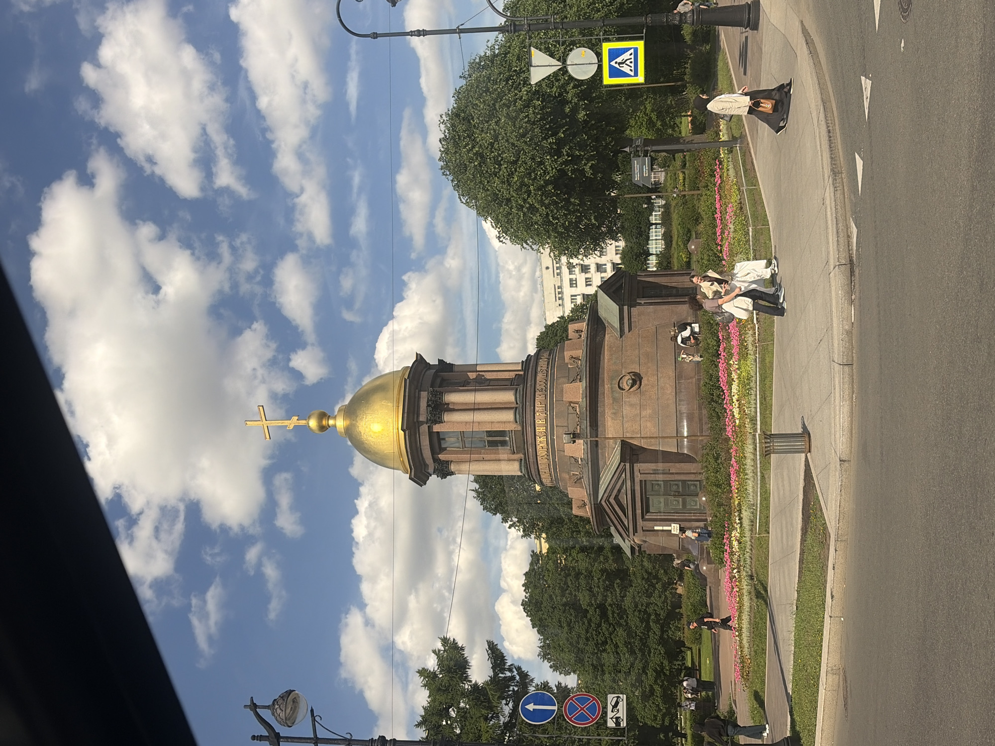 The image shows a view of the Naval Cathedral of Saint Nicholas in Kronstadt, Saint Petersburg, Russia, on a sunny day with scattered clouds. The domed cathedral, with its golden cross, is the central focus. A lamppost is visible to the left of the cathedral, and a tree is to its left and right. Several traffic signs are present, including a blue circular sign with a white arrow pointing up, a red circular sign with a diagonal red bar, and a yellow square sign with a blue pedestrian crossing symbol. In the foreground, a sidewalk curves around a circular flower bed filled with pink and white flowers. Several people are walking on the sidewalk and in the street. Some people are dressed in casual clothing, while others are wearing hijabs. The street is paved and appears to be a busy urban area.