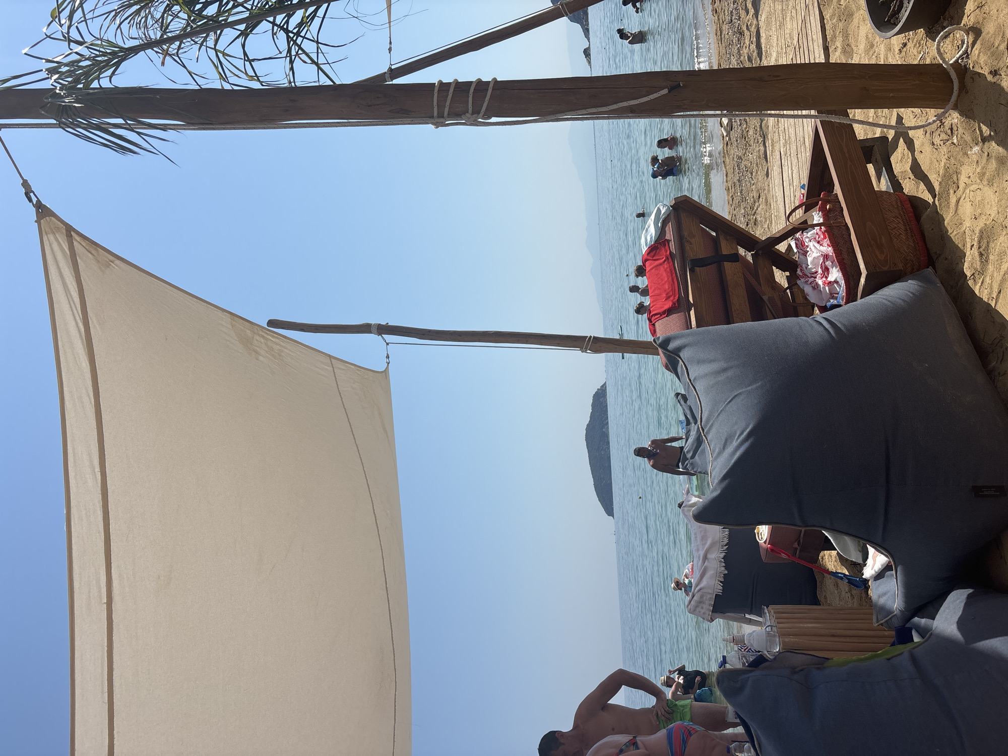 A bright, sunny day at the beach in Nea Iraklitsa, Greece. The foreground is dominated by two large, dark blue beanbag chairs on a sandy surface. Behind the chairs, a wooden beach lounger is set up, holding a red towel and a basket filled with white and red patterned fabric. A wooden support pole with rope detailing stands to the right of the lounger, part of a canopy structure that extends overhead, providing shade.
In the middle ground, several people are visible in the shallow, calm, turquoise water. Some are standing, others are interacting. A small, distant island with green foliage is visible on the horizon. The sky is a clear, bright blue. The overall impression is of a relaxed, recreational beach environment.