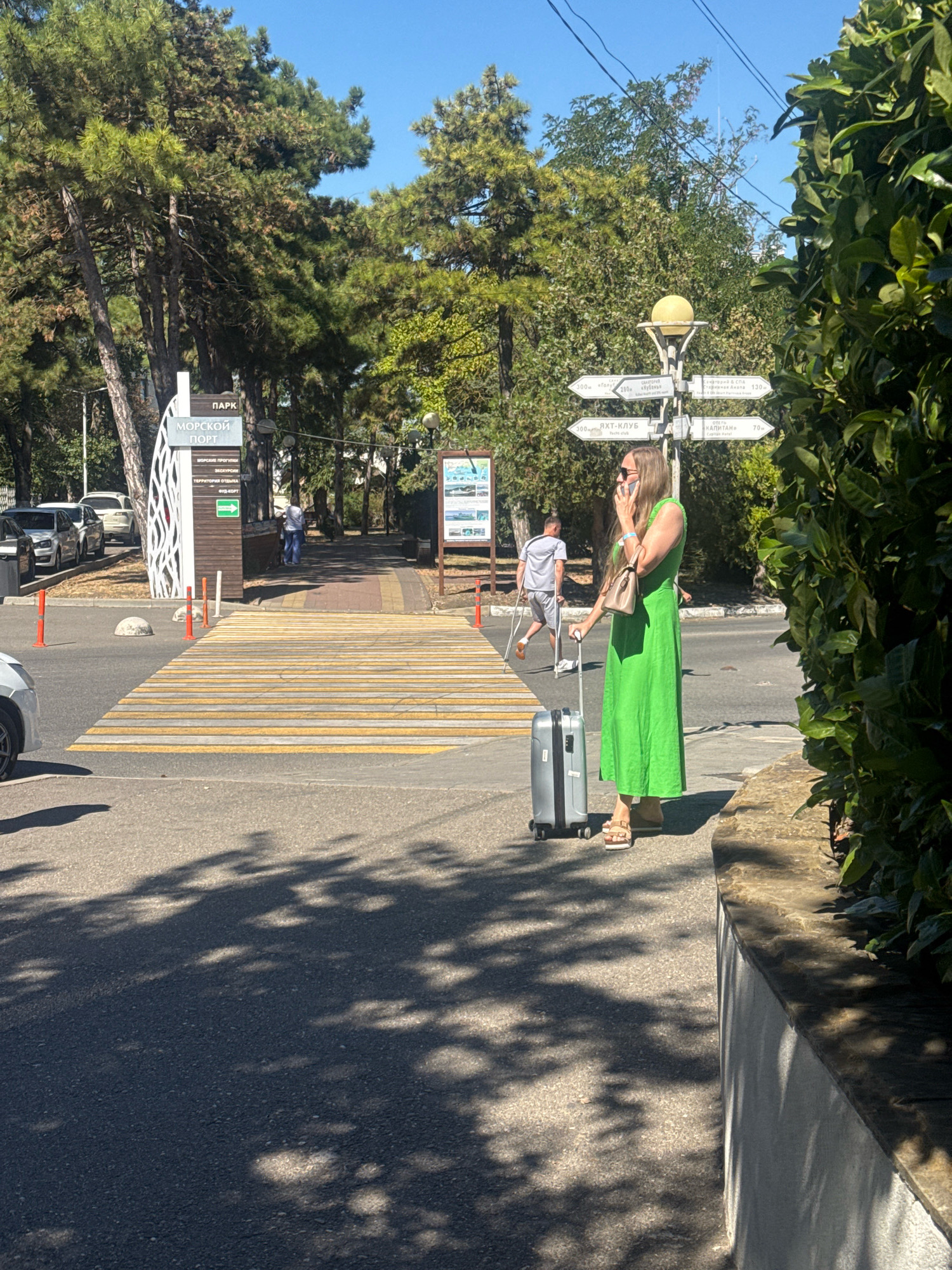 The image depicts a sunny day scene in Anapa, Russia, likely near a park or promenade. A woman in a bright green dress stands on a sidewalk, holding a phone to her ear and a small beige purse. She is next to a silver rolling suitcase. In the background, a pedestrian crossing with yellow and white stripes is visible. Behind the crossing, a signpost indicates directions to "PARK," "SEA PORT," "SEA CRUISES," "RECREATION AREA," and "PRIVATE BEACH." Further in the background, several people are walking, and trees line the path. A street lamp with a yellow globe stands at the intersection of several directional signs. The overall environment suggests a tourist area, possibly a resort town, with lush greenery and clear blue skies.