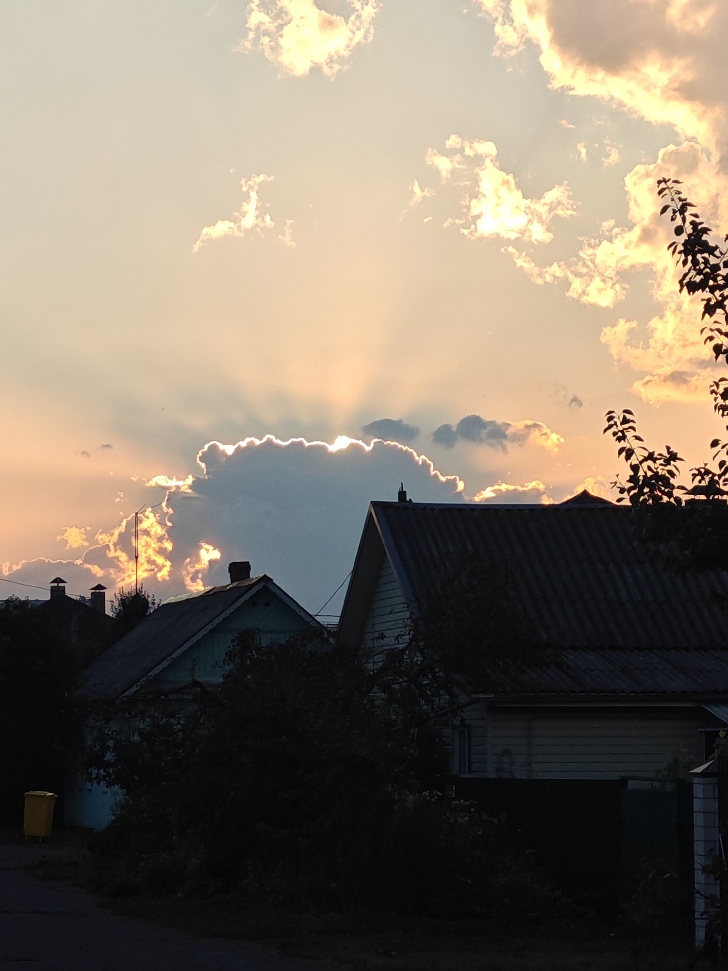 The image depicts a sunset sky over a residential area in Homyel, Belarus. The sun is partially obscured by a large, fluffy cloud, casting rays of golden light through the sky. In the foreground, silhouetted against the bright sky, are the dark shapes of several houses and trees. The houses have sloped roofs, and some have chimneys visible. A yellow garbage bin is visible on the left side of the image, near a dark, possibly asphalt road. The overall impression is of a peaceful, rural evening scene.