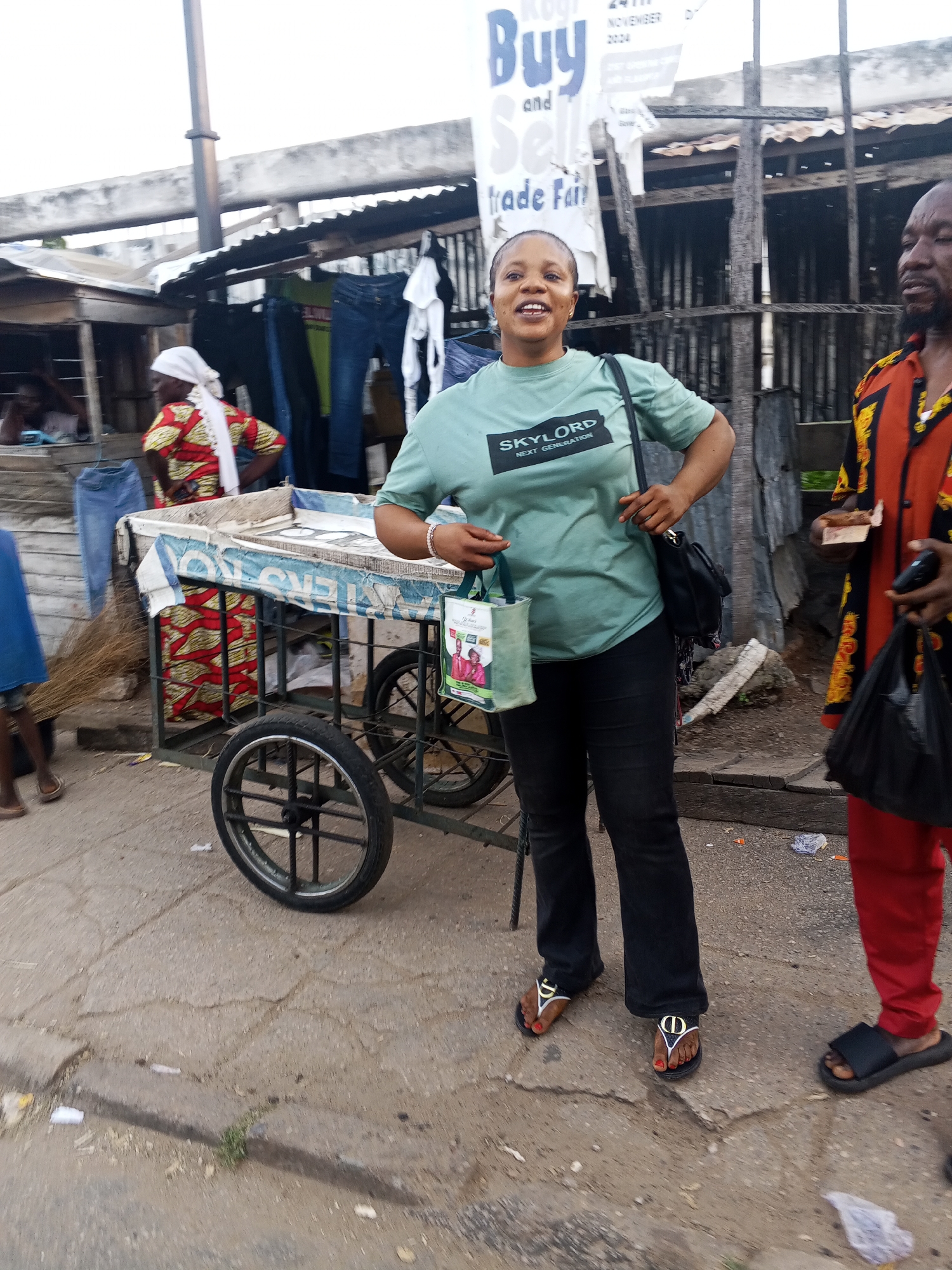 A woman stands with a large shopping cart full of items in an outdoor market. The woman, wearing a green t-shirt and black pants, smiles at the camera while holding a reusable shopping bag. Behind her, a man in an orange and black patterned shirt looks away from the camera. A large banner with the words "Buy and Sell Trade Fair" is visible in the background. The ground is paved with cracked concrete, and there are various stalls and structures that appear to be made of wood and corrugated metal. The overall scene suggests a bustling marketplace environment.