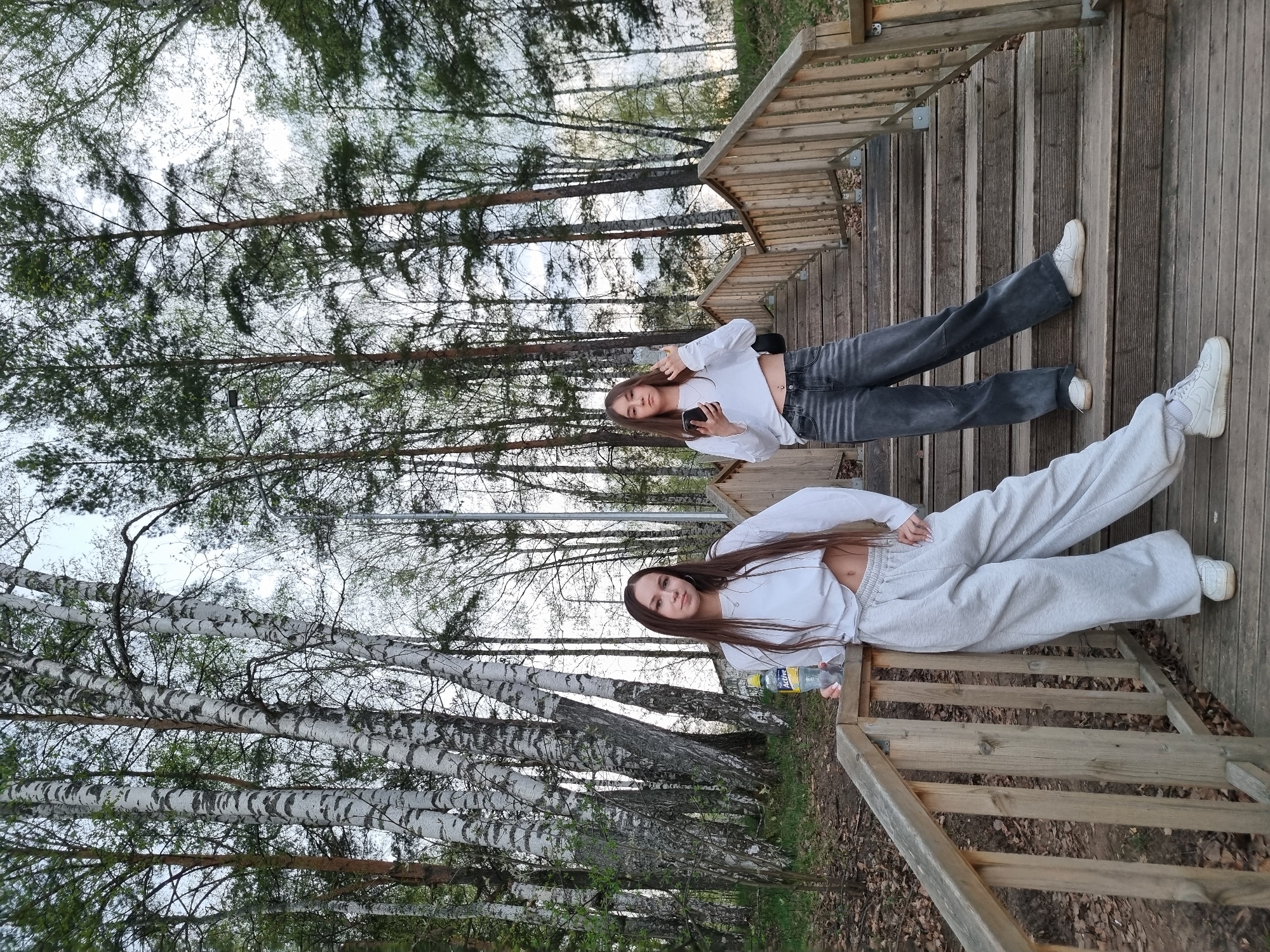 Two young women are lying on wooden stairs outdoors. The woman in the foreground wears light gray sweatpants and a white cropped long-sleeved shirt, holding a bottle of water. The woman in the background wears gray jeans and a white cropped long-sleeved shirt, holding a cell phone. The scene appears to be in a park or wooded area, with birch trees and other trees visible in the background. It is likely the city of Chaikovsky, Russia, based on the prompt.