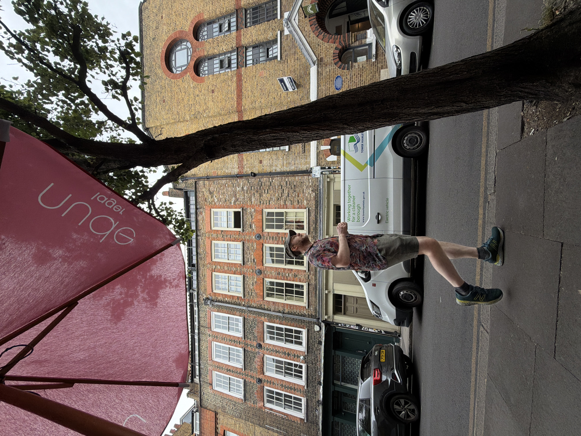 The image captures an outdoor street scene in London, United Kingdom. A man wearing a patterned shirt, khaki shorts, a cap, and green athletic shoes is standing on the sidewalk. He appears to be looking upwards. Behind him are buildings of brick construction. The building on the left features numerous rectangular windows, while the building on the right has unique circular windows near the top. There's a white commercial van with green and blue accents and the words "TOWER HAMLET" on its side parked on the street, as well as a dark-colored car with a license plate reading "LP24 GXN." A tree trunk and a partially visible red umbrella with a word that appears to be "Lunge" in reverse, also in white, frame the upper and left parts of the image. A double yellow line is painted on the edge of the street. A green sign reads "LET" over a phone number, "020 7749 7650." "Electric vehicle" is also written on the van.
