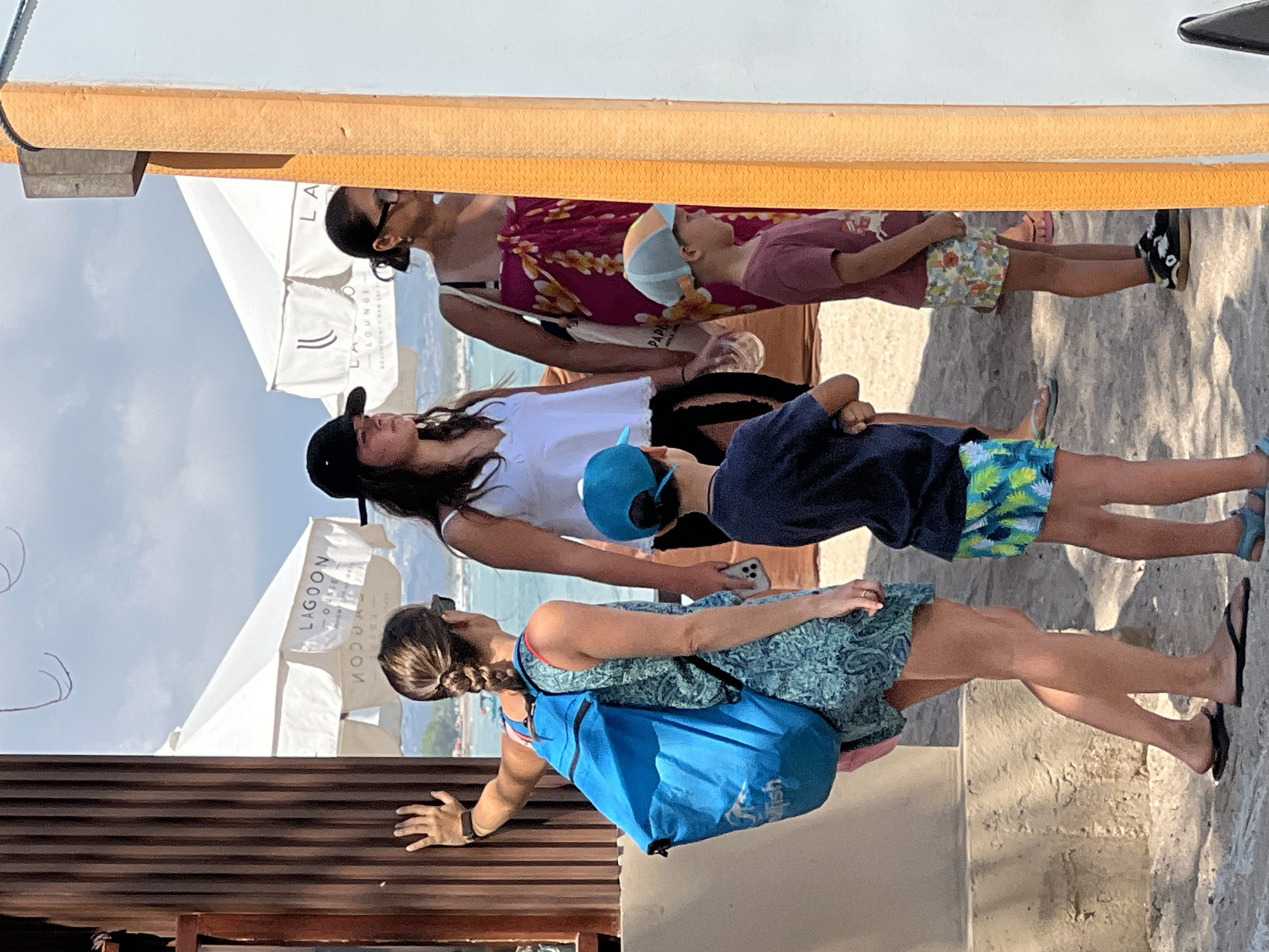 The image captures a group of people on a sandy beach with ocean and sky in the background. In the foreground, a woman with braided brown hair wearing a blue patterned dress and carrying a blue backpack stands on the left. To her right, a person in a white tank top and black cap is visible, followed by another woman in a pink sarong with a floral print. Two young boys are also present; one in a blue cap and dark shirt with colorful shorts, and another in a maroon shirt and floral shorts. Beach umbrellas are set up behind the individuals. The scene appears to be at a beachside establishment in Kuta, Indonesia, indicated by the "Lagoon Lounge Beachfront Bar and Spa" signage on an umbrella. The lighting suggests it is daytime, likely mid-morning or afternoon.