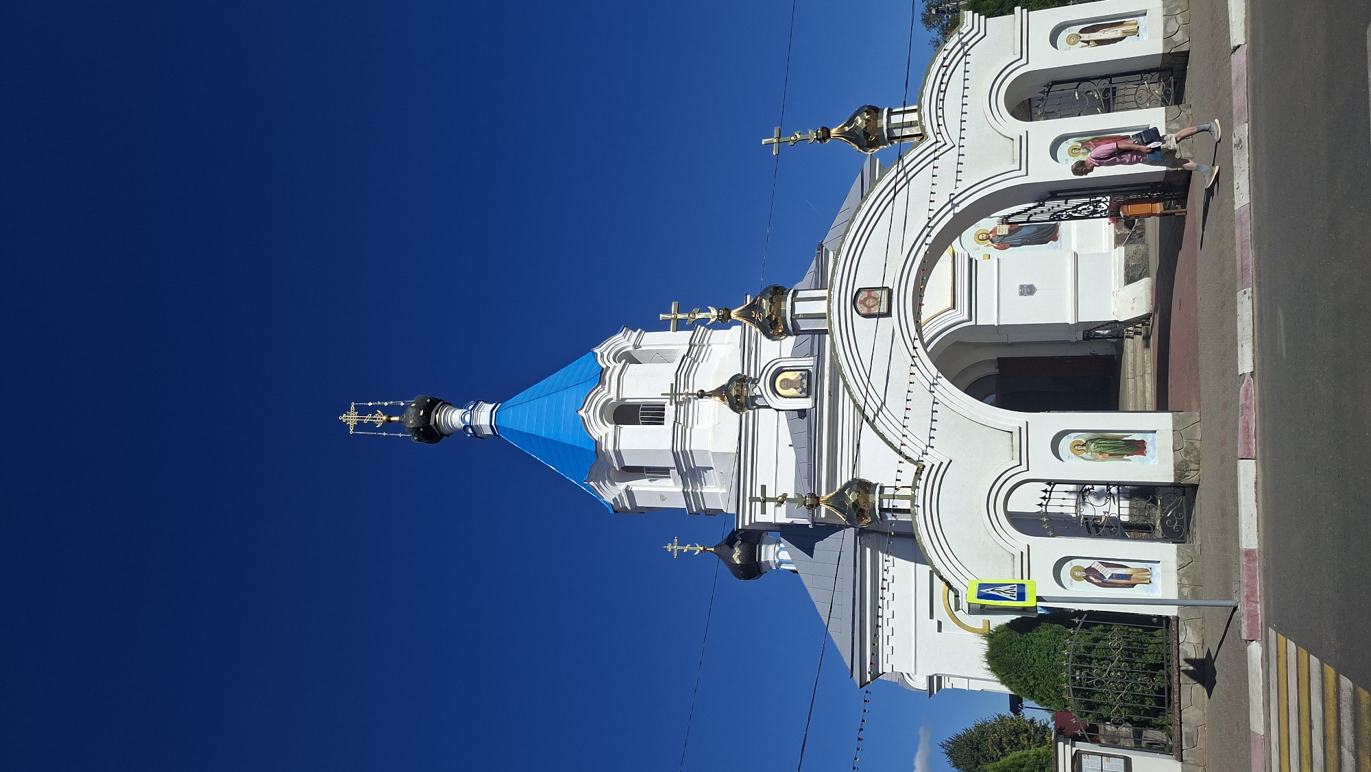 The image shows a white Orthodox church with a bright blue dome and gold-plated cupolas. It is a sunny day with a clear blue sky. The church has a large arched entrance with a black metal gate. There are religious icons painted on the exterior walls of the church, near the entrance. A pedestrian crossing with yellow and white stripes is visible in the foreground. A yellow and blue pedestrian crossing sign is mounted on a pole to the left of the entrance. A person, wearing a pink shirt and grey shorts, is walking on the sidewalk in front of the church, carrying a dark bag. Trees and a black metal fence are visible to the left of the church. The church is located in the city of Pastavy, Belarus.