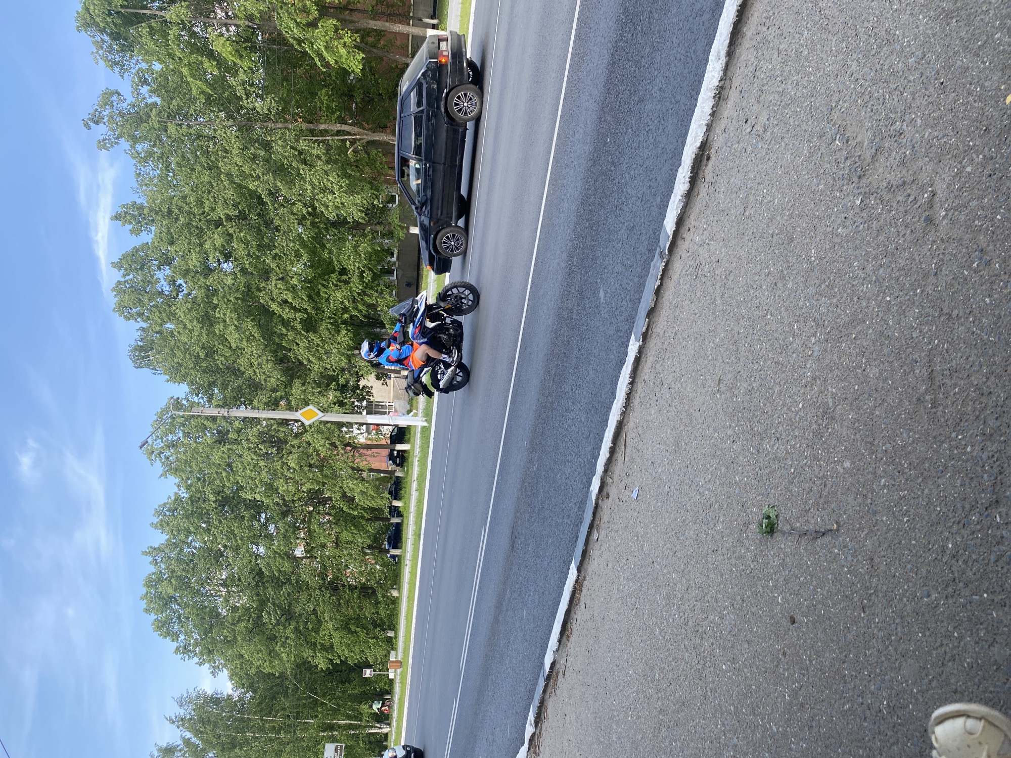 The image depicts a street scene in what appears to be the city of Chaikovsky, Russia.
**Setting:** The location is on the side of a road with an adjacent sidewalk. Lush green trees line the road, suggesting a residential or suburban area. The sky is blue with some light clouds.
**People:** A person wearing a helmet and brightly colored riding gear (orange and blue) is riding a blue and silver motorcycle on the road.
**Objects:** A black car is parked on the side of the road. There are road markings, a street sign, and what appears to be litter on the sidewalk.
**Actions:** The motorcyclist is driving on the road. The car is parked.
**Environment:** The environment is a sunny day with a tree-lined road and a sidewalk.