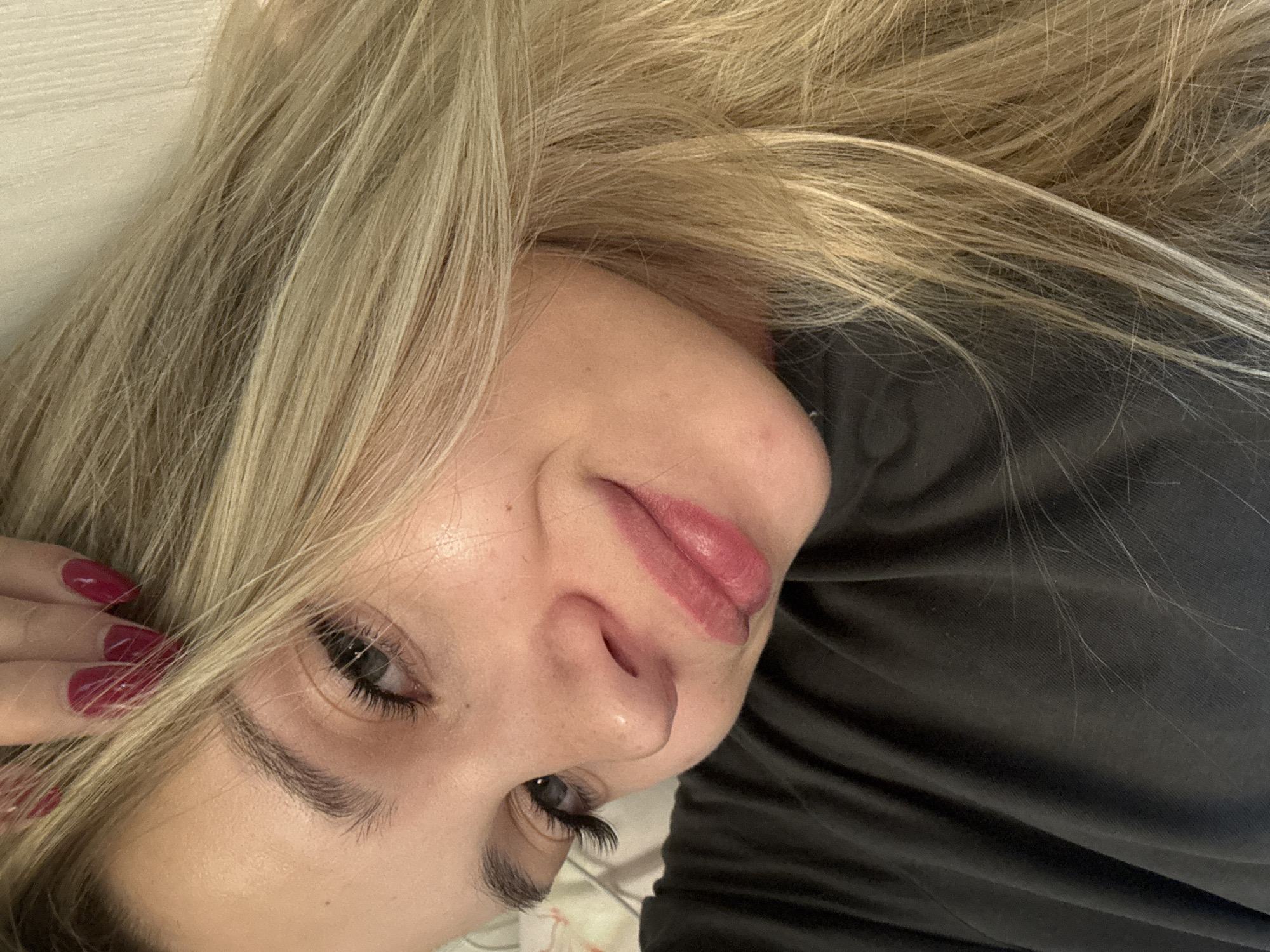 The image is a close-up selfie of a young woman with blonde hair. She is wearing a dark t-shirt. Her left hand, with dark red nail polish, is resting on top of her head and hair. The woman is looking towards the camera with a slight smile. The background is out of focus and appears to be indoors, possibly a bed. The image does not contain any information about the location in the city of Chaikovsky, Russia.