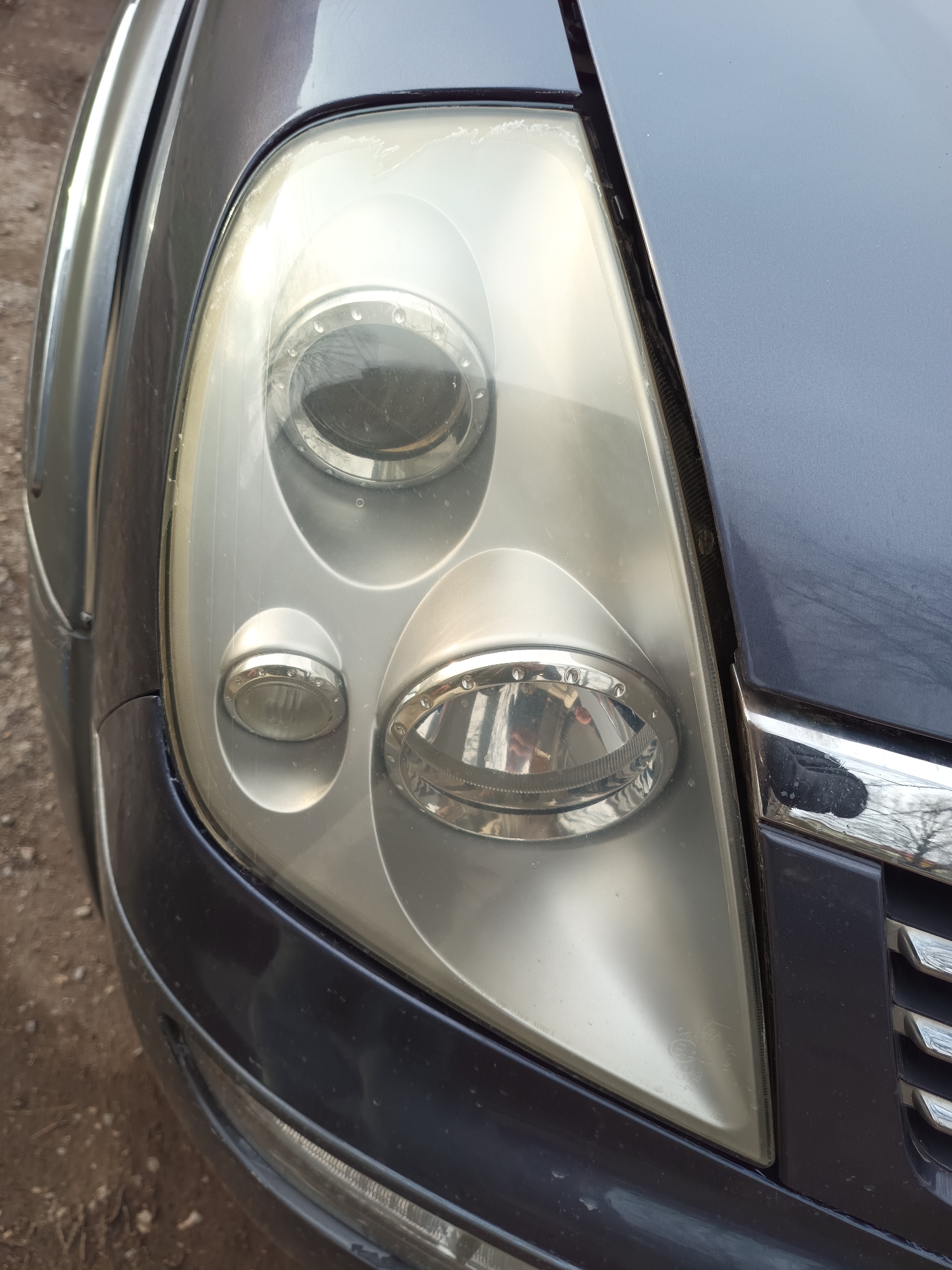 The image shows a close-up of the front right headlight of a dark blue or grey car. The headlight lens is cloudy or faded, obscuring the internal components of the light. The car's body paint appears metallic. The background is out of focus, revealing dirt or gravel on the ground and hints of vegetation. There's no clear indication of the image being taken in Targovishte, Bulgaria, or any specific urban location.
