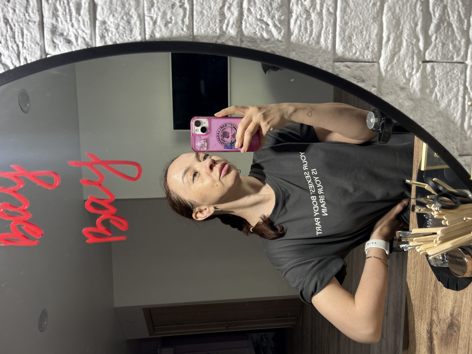 A woman is taking a selfie in a mirror. The mirror has red lettering on it. She is holding a pink phone. She is wearing a black t-shirt with white lettering. There is a small heart tattoo on her arm. Her hair is in a braid. In the mirror is a white brick wall and a black rectangle. To the right of the woman in the image is a wooden table with various items on it, including what appears to be a set of makeup brushes and containers. The location appears to be Chaikovsky, Russia.