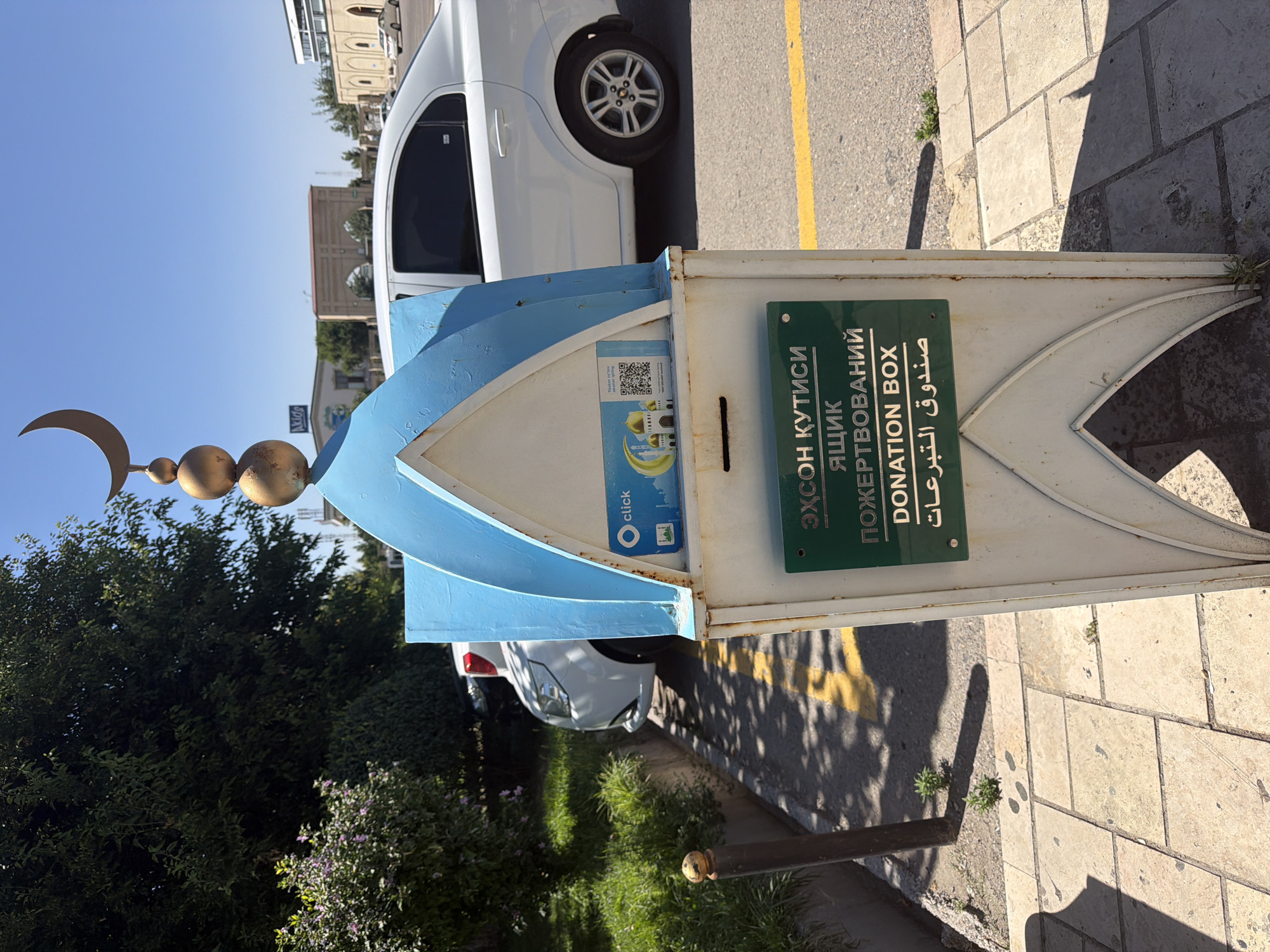 A donation box, designed to resemble a mosque with a blue domed roof and a crescent moon on top, stands on a paved sidewalk next to a street. The box is white with some rust, and it features green signage in Cyrillic and English stating "DONATION BOX." A QR code and the "Click" logo are visible on the upper portion of the box. A white car is parked on the street behind the donation box, and a lush green tree with some flowering bushes is to the left. The scene appears to be in Zangiata, Uzbekistan, suggested by the architectural style and the Cyrillic script. The sky is clear and blue, indicating a sunny day.
