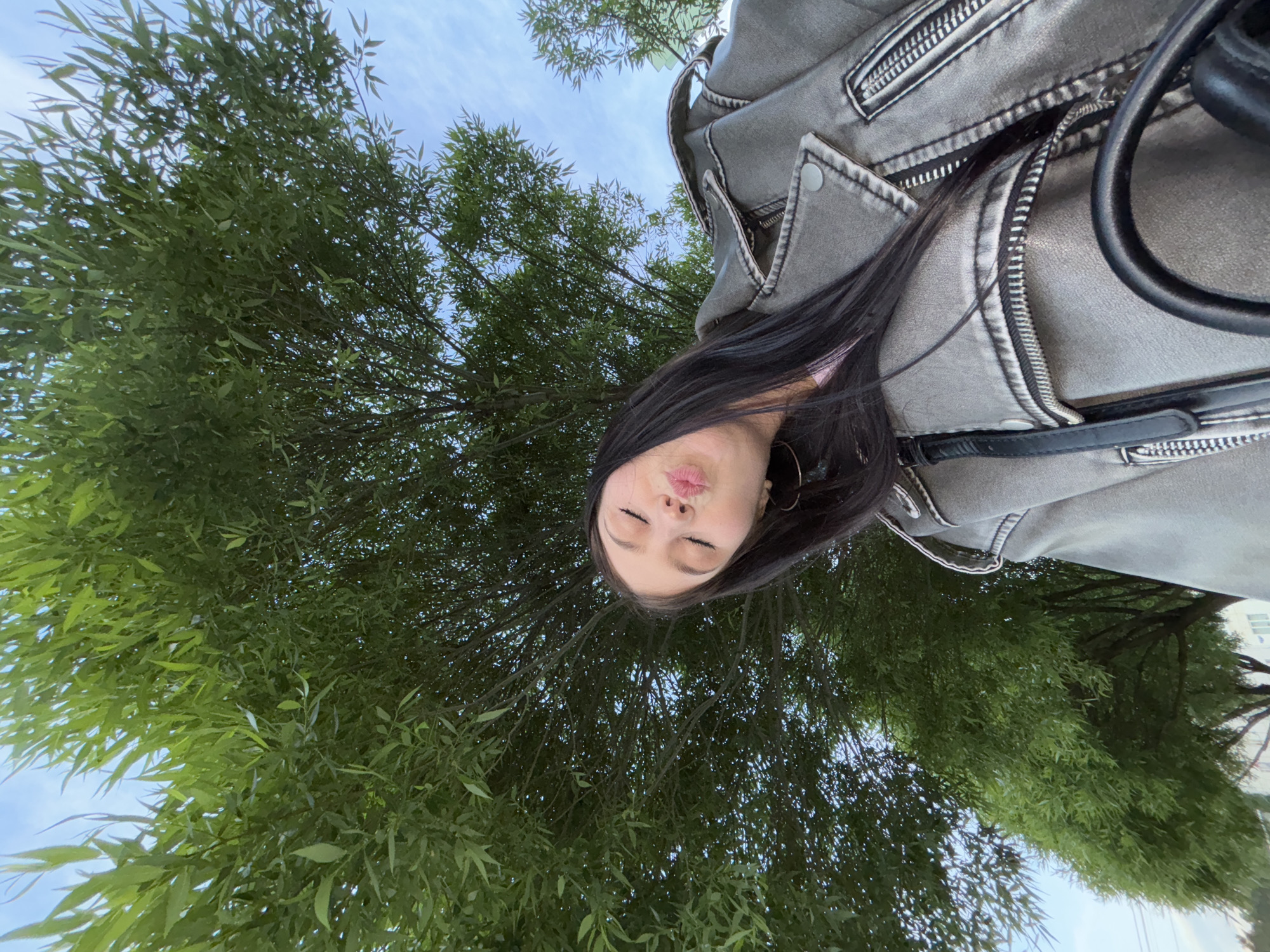 A low-angle shot captures a woman with long dark hair puckering her lips toward the camera. She is wearing a gray jacket with zipper details. Behind her, a lush green tree fills much of the frame, partially obscuring a light blue sky. The location is Chaikovsky, Russia.