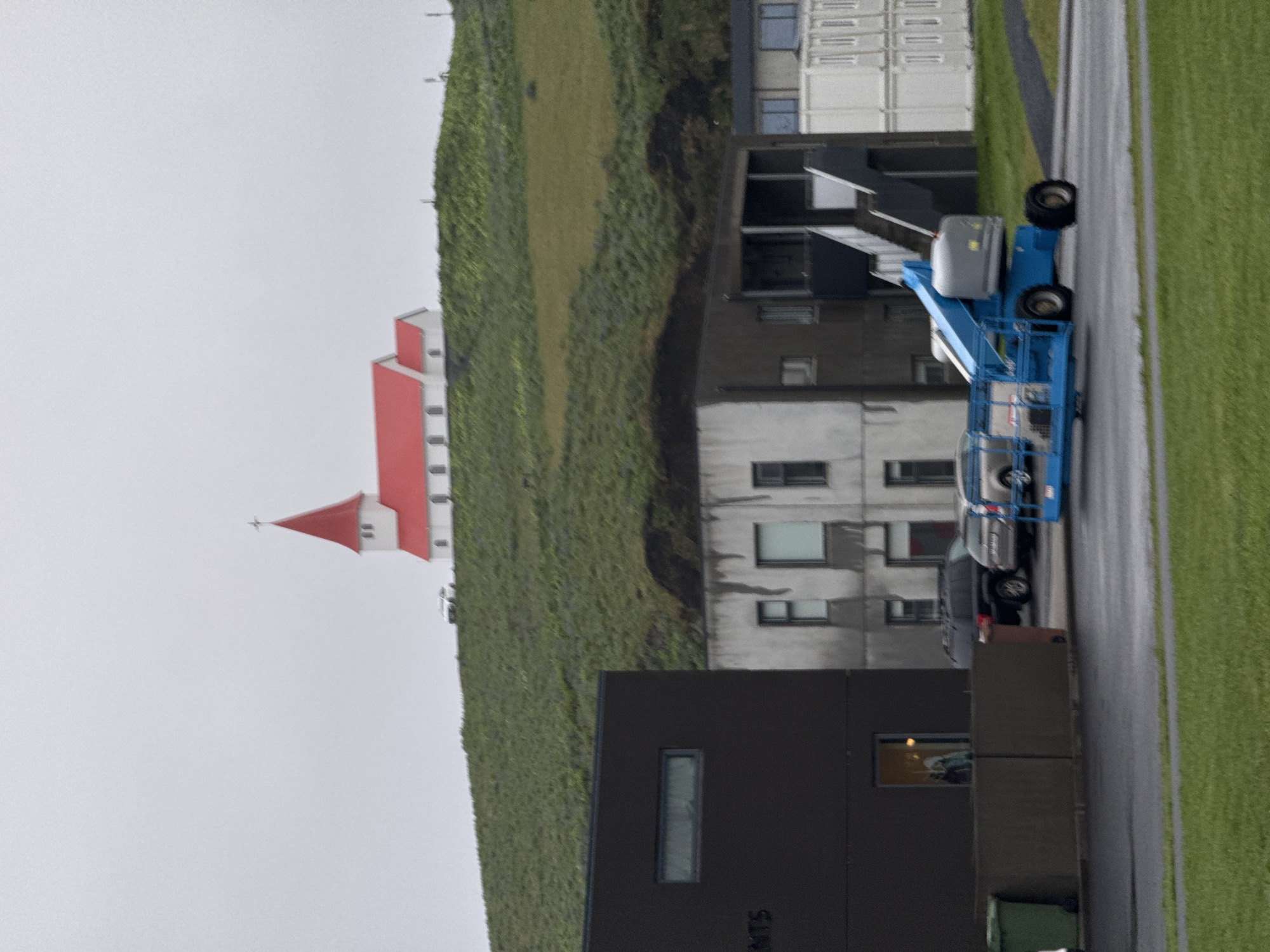 The image shows a scene in Vik, Iceland. A church with a red roof and white walls is situated on a green hillside. Modern buildings are near the church, including a dark-colored building and a gray building with rectangular windows. A blue boom lift is parked on the asphalt road near the buildings. Green grass covers a portion of the roadside, and a cloudy sky is overhead.