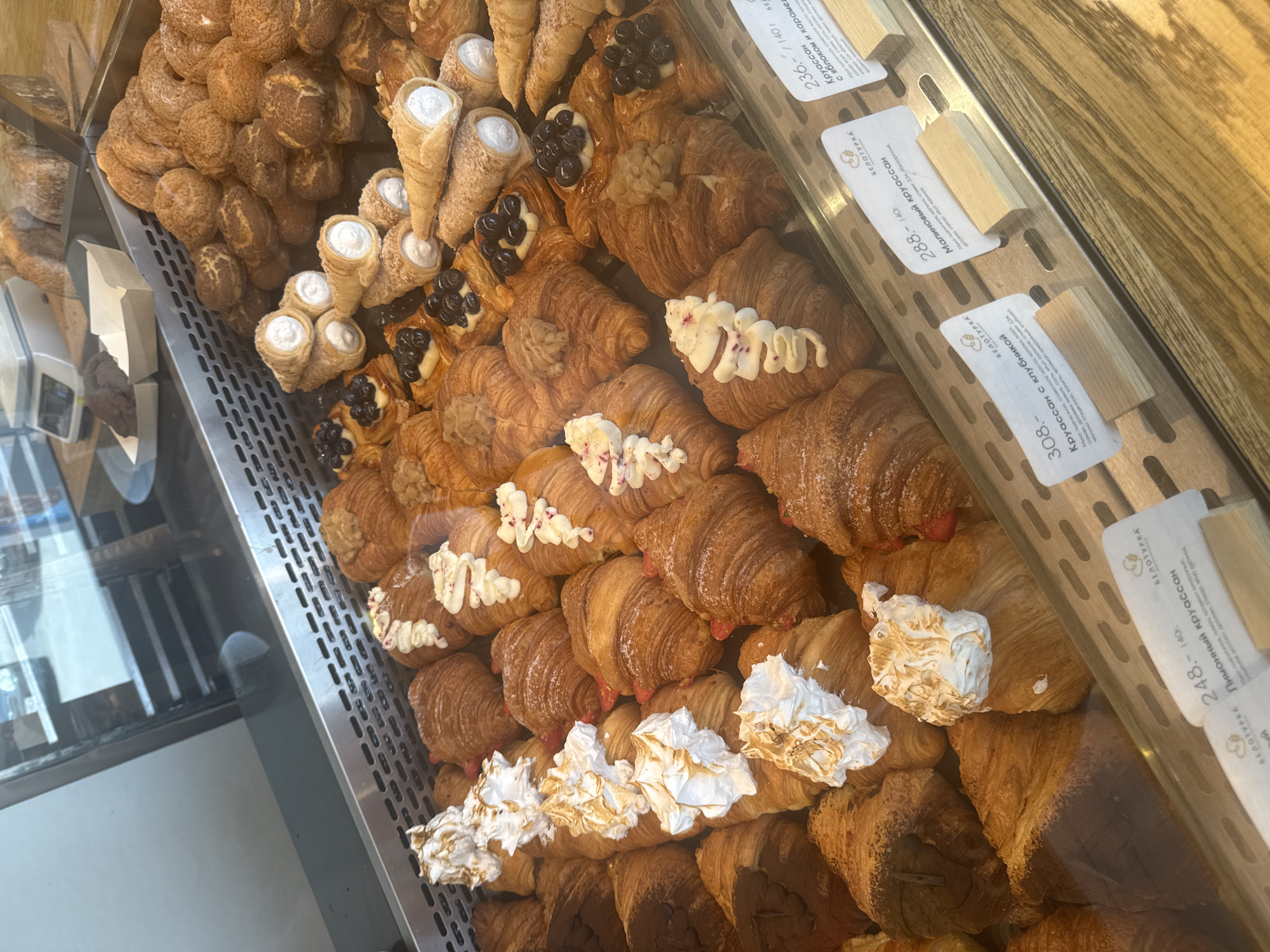 The image depicts a bakery display case filled with a variety of pastries. On the left, there's a pile of round, textured pastries, next to cone-shaped pastries filled with white cream. Adjacent to these are croissants topped with dark berries and cream. The majority of the display consists of golden-brown croissants with various toppings: some have white icing, others have a reddish filling, and some are topped with meringue. Price tags with Russian text are visible along the edge of the display case. Based on the Russian text on the price tags, the bakery could be in Izhevsk, Russia.
