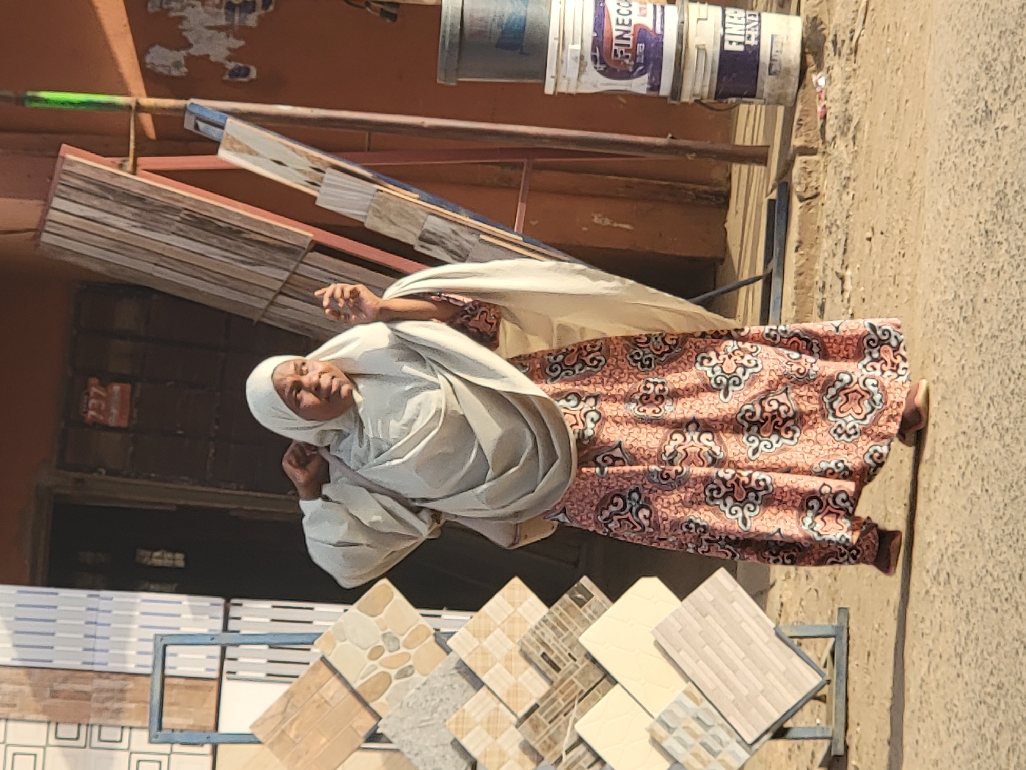 The image depicts an elderly woman standing in front of a tile shop in Maiburiji, Nigeria. She is wearing a patterned dress and a white headscarf. In front of her, tiles are arranged in a display stand. Building materials are visible nearby, including stacks of wooden planks and buckets of paint. The street appears to be unpaved.
