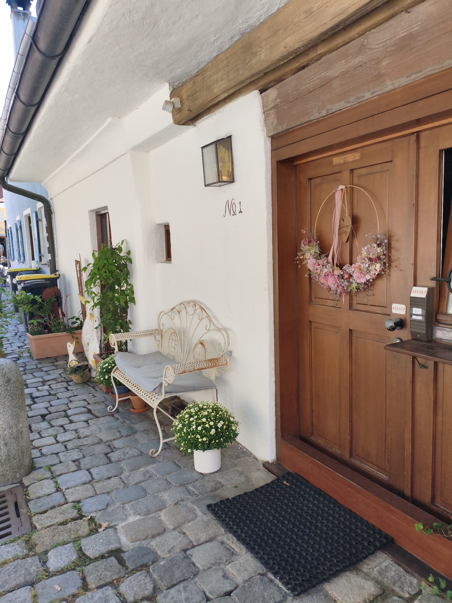 The image shows a charming, narrow cobblestone alleyway in Landsberg am Lech, Germany. On the right, a weathered wooden double door serves as the entrance to a building. A festive wreath adorned with pink flowers and greenery hangs on the door. A small black doormat is placed on the cobblestones in front of the entrance. To the left of the door, a white metal bench with a grey cushion is placed next to a potted white chrysanthemum. Further to the left, several plants in terra cotta pots line the white-washed wall of the building. Decorative garden ornaments, including a duck, are also visible among the plants. The cobblestone path leads further into the alley, with trash bins and more buildings visible in the background. The scene evokes a quiet, picturesque European street.