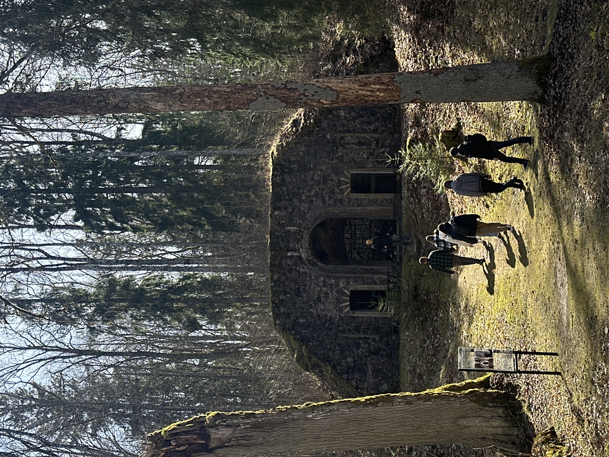 The image shows a stone building in a wooded area near Bīriņi, Latvia. The building has an arched entryway with a barred gate and two windows on either side of the entrance. The structure appears to be moss-covered and constructed from rough-hewn stones. A group of people are walking on a path adjacent to the building, and their shadows are visible on the ground. One person is standing near the entrance. The surroundings consist of trees and ground covered with grass, moss, and fallen leaves. A sign or display stand is placed on the edge of the path. The time of day is likely daytime, with sun shining through the trees, creating shadows and highlights.