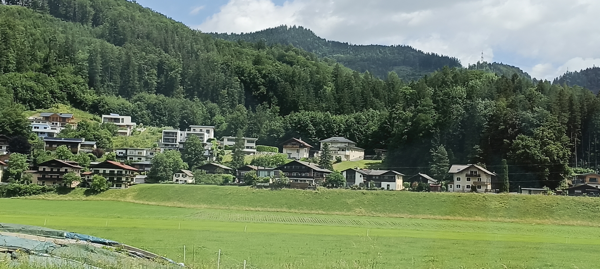 The image captures a scenic view of the city of Torren, Austria, nestled at the base of a verdant, tree-covered mountain. A cluster of houses, characterized by their varied architectural styles and roof colors, are built into the hillside, suggesting a mountainous terrain. The houses range from modern, multi-story structures with large windows to more traditional chalets. In the foreground, a lush green field stretches across the landscape, with faint rows indicating agricultural activity. The sky above is partly cloudy, with patches of blue visible between the white clouds. The overall impression is one of a tranquil, European alpine village.
