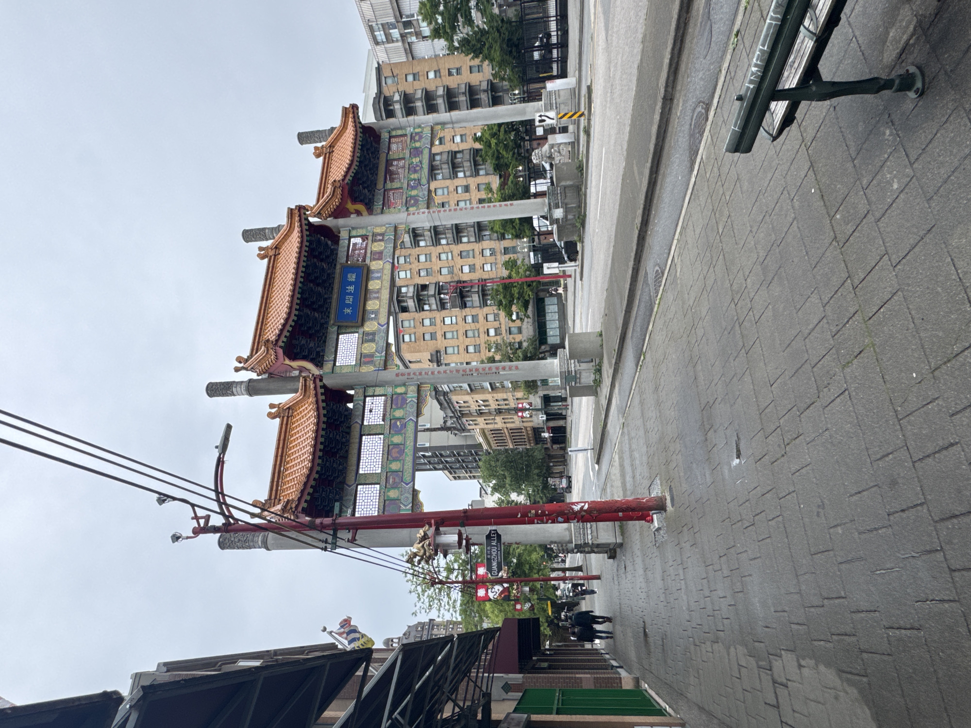 The image depicts the entrance to Chinatown in Vancouver, Canada, on a cloudy day. A large, ornate archway with traditional Chinese architecture, featuring red and gold roofs, spans across a street. The archway is supported by pillars adorned with intricate carvings and colorful patterns. A blue sign with Chinese characters is visible on the archway.
To the left of the archway, a brick building with awnings lines a sidewalk. The sidewalk is paved with grey stone tiles and has a dark-colored awning covering shops. Several people are walking on the sidewalk, some dressed in dark clothing. A red lamppost with decorative elements stands prominently on the sidewalk. Street signs, including one indicating "Guangzhou Alley," are attached to the lamppost.
The street itself is paved and appears to be relatively empty, with a few cars parked or driving in the distance. Buildings of varying heights and architectural styles are visible on the opposite side of the street, including apartment buildings. In the foreground, a dark green park bench with wooden slats is situated on the sidewalk. The overall environment suggests an urban setting with a strong cultural presence.