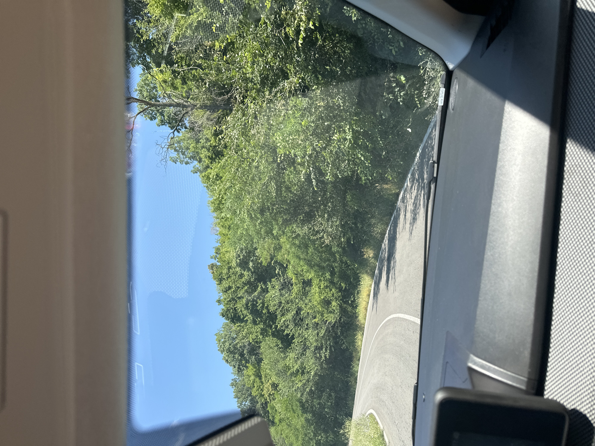 The image shows a view from inside a car looking out through the windshield onto a winding asphalt road bordered by dense green trees. The sky is clear and blue. A black device, possibly a dashcam, is mounted on the car's dashboard in the lower left corner. The road curves to the left, with white lines indicating the lanes. The environment suggests a rural or natural setting, possibly in or around Karnare, Bulgaria, given the terrain and vegetation. The lighting indicates it is daytime.