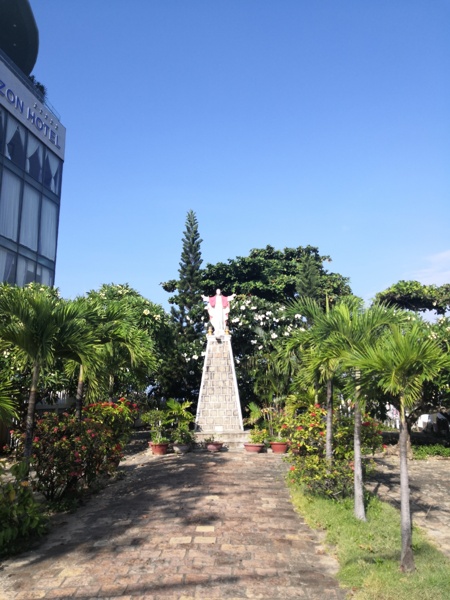 The image displays an outdoor, landscaped scene under a clear blue sky in Bac Nha Trang Ward, Vietnam. In the upper left, a modern, multi-story hotel building is visible, featuring a sign that reads "ZON HOTEL" with four stars. The building has a dark, rounded structure on its roof and large, rectangular windows. A paved walkway, constructed from light-colored, irregular pavers, extends from the foreground towards a central statue. This statue, depicting a white figure with outstretched arms and a red garment, stands atop a tall, narrow pedestal made of light-colored, rectangular stone blocks.
The area is densely vegetated with tropical plants. Several palm trees with slender, light-colored trunks are positioned along the paved path, particularly on the right side. Bushes bearing small red flowers are abundant along the walkway. Behind the statue and other foliage, large trees with white flowers (likely plumeria) and a tall, slender conifer-like tree are visible. Potted plants are arranged around the base of the statue's pedestal and along the path. Sunlight casts distinct shadows from the trees across the paved walkway. No individuals are present in the depicted scene.