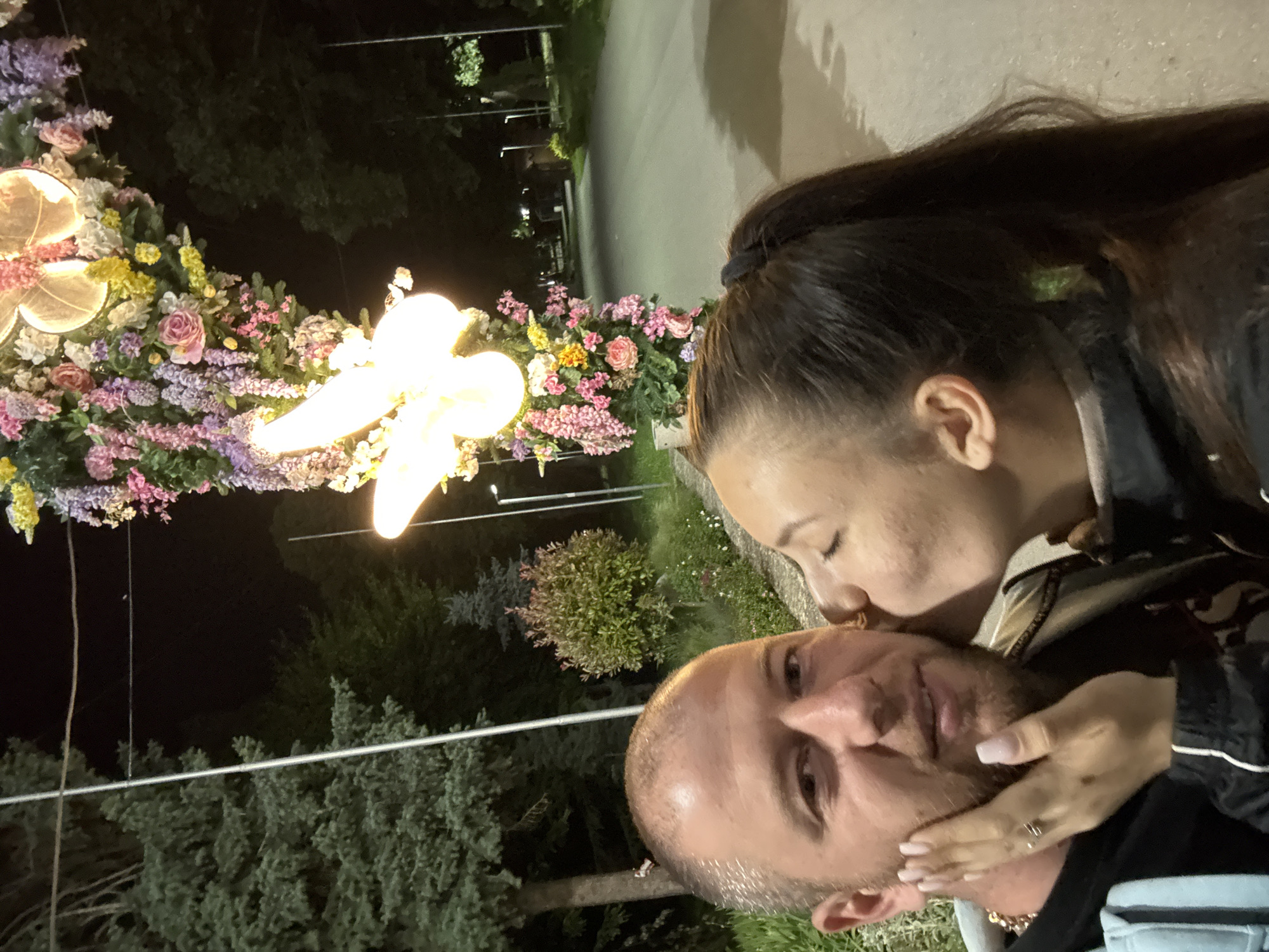 The image shows a man and a woman in an embrace. The woman is kissing the man on the cheek. The man is looking directly at the camera and has a short, closely-cut haircut and light stubble. The woman has her hair pulled back in a high ponytail, and one hand is touching the man's face. They appear to be standing outdoors at night.
Behind them is a large floral display illuminated with lights. The flowers include roses and other colorful blooms. In the background, there are trees and what appears to be a path or road with lampposts.
Based on the description and other visual cues, the photograph likely captures a moment near the Shumen Municipality building. The flower display likely is related to public events and the surrounding environment, which includes city infrastructure and vegetation.