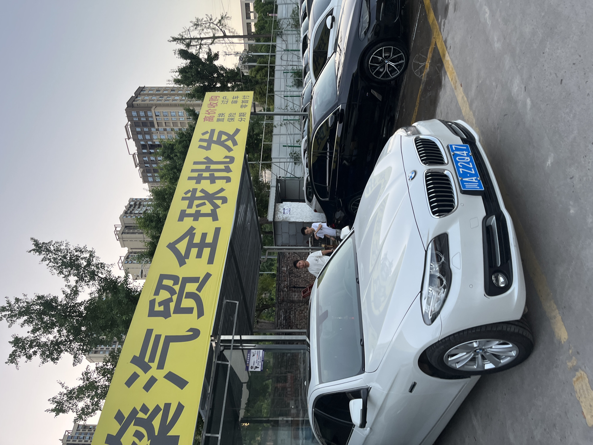 The image shows a car lot in Banan, China, with several parked cars and a large yellow sign with Chinese characters. The foreground features a white BMW with a blue license plate reading "A-Z2047". Behind the BMW are other cars, including a black car with visible wheel details. The sign reads "Mei Qi Mao Quan Qiu Pi Fa" vertically. Two people are standing near the back of the car, partially obscured by the sign. The background includes buildings, trees, and a partial view of the sky. The ground appears to be a paved parking area with yellow parking lines.