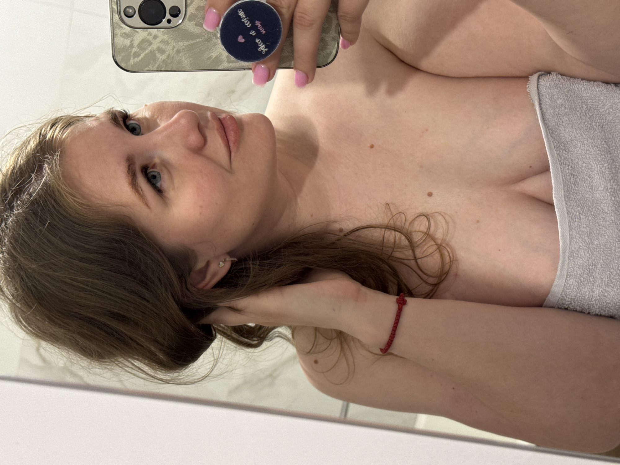 A female individual with light skin, brown hair, and blue eyes is captured in a mirror selfie within a bathroom in Ферма, Russia. She is wrapped in a light grey terrycloth towel, covering her chest and torso. Her left hand is raised to her right ear, appearing to adjust or hold her hair. In her right hand, she holds a smartphone (an iPhone model with multiple rear cameras) that has a textured, light-colored case and a dark blue PopSocket featuring a pink heart icon and mirrored white text. Her nails are painted a light pink. A red knotted string bracelet is worn on her left wrist, and she has a small silver heart stud and a small silver hoop earring in her right ear. Several small moles are visible on her neck, shoulders, and upper chest. The background shows plain, light-colored tiled walls, characteristic of a bathroom, reflected in the mirror.