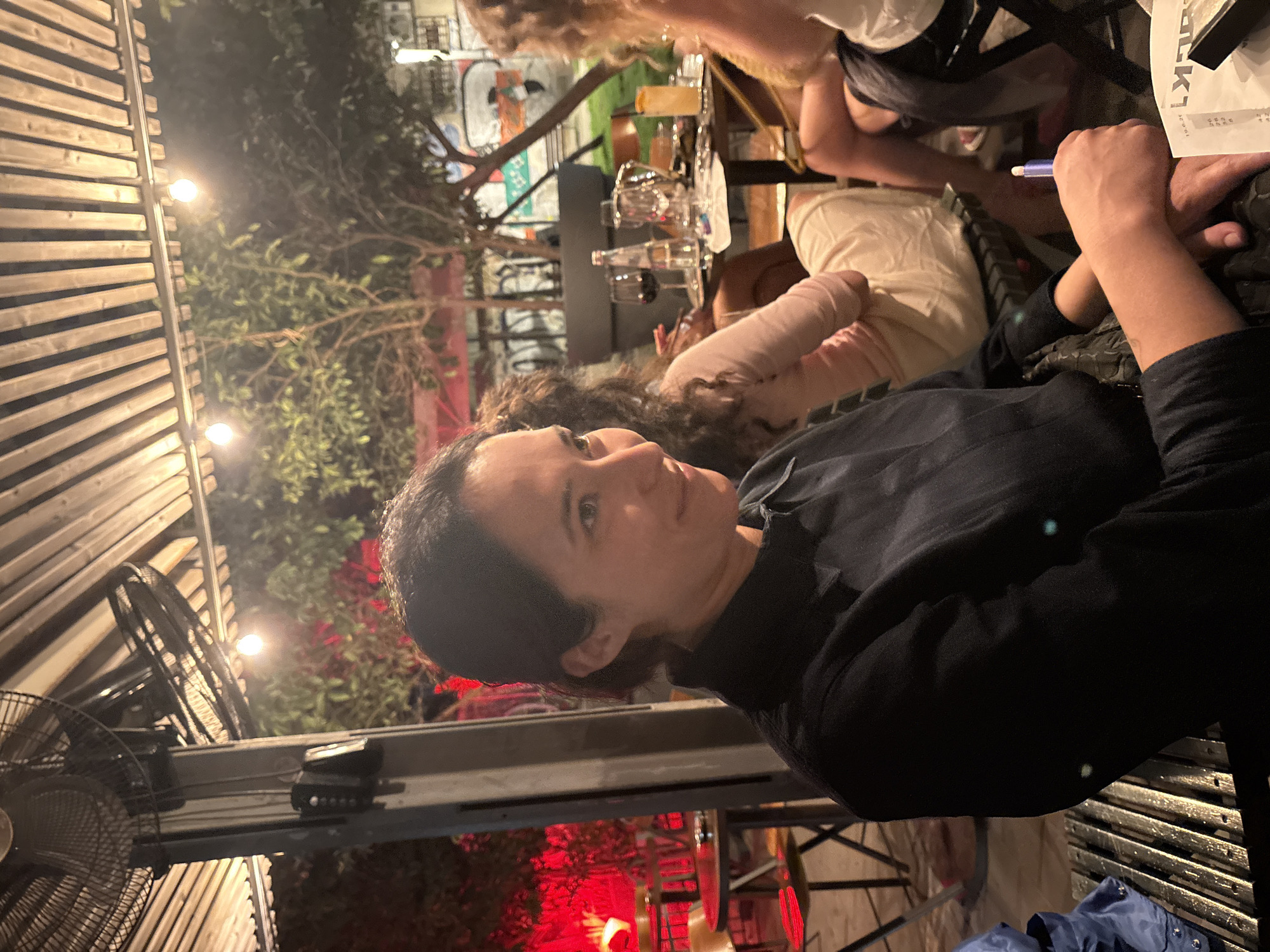 A medium shot captures a woman in the foreground, seated at an outdoor table. She is wearing a dark collared shirt. Her head is turned to her left, and she is looking slightly downwards and to her left, with a subtle smile. Her dark hair is pulled back. Behind her, at the same table, are other individuals, including a woman with curly hair wearing a light-colored top, and partially visible individuals further back. The table is set with drinks, including wine glasses and a cocktail. The setting appears to be a casual outdoor dining area, possibly a restaurant or bar, with a slatted roof overhead, string lights, and greenery in the background. Red lighting emanates from behind some tables on the left side of the image, creating a dramatic effect. Graffiti is visible on a wall in the far background. The scene is likely in Tel Aviv, Israel, given the presence of outdoor dining and the general ambiance.