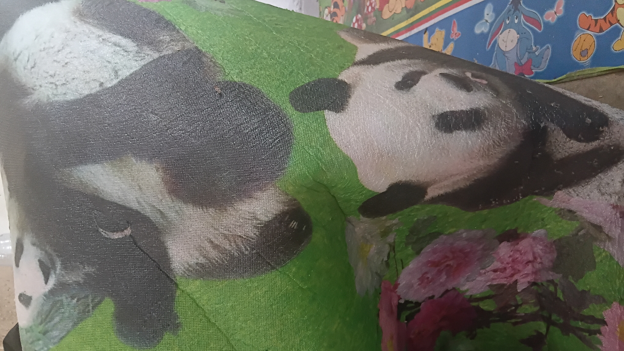 The image displays a close-up, slightly blurry view of a fabric item, possibly a bag or banner, featuring a stylized depiction of pandas and flowers. The fabric has a textured surface with a visible weave pattern. In the foreground, pink and white flowers are visible against a green background, with parts of a panda's face and body also discernible. The composition is dynamic, with the fabric appearing to be draped or folded, creating curves and shadows. In the background, a portion of a mat with cartoon characters, including Winnie the Pooh and Tigger, is visible. The lighting suggests an indoor setting with overhead illumination, casting subtle highlights and shadows on the fabric. No elements in the image suggest a specific location within the city of Prabumulih, Indonesia.